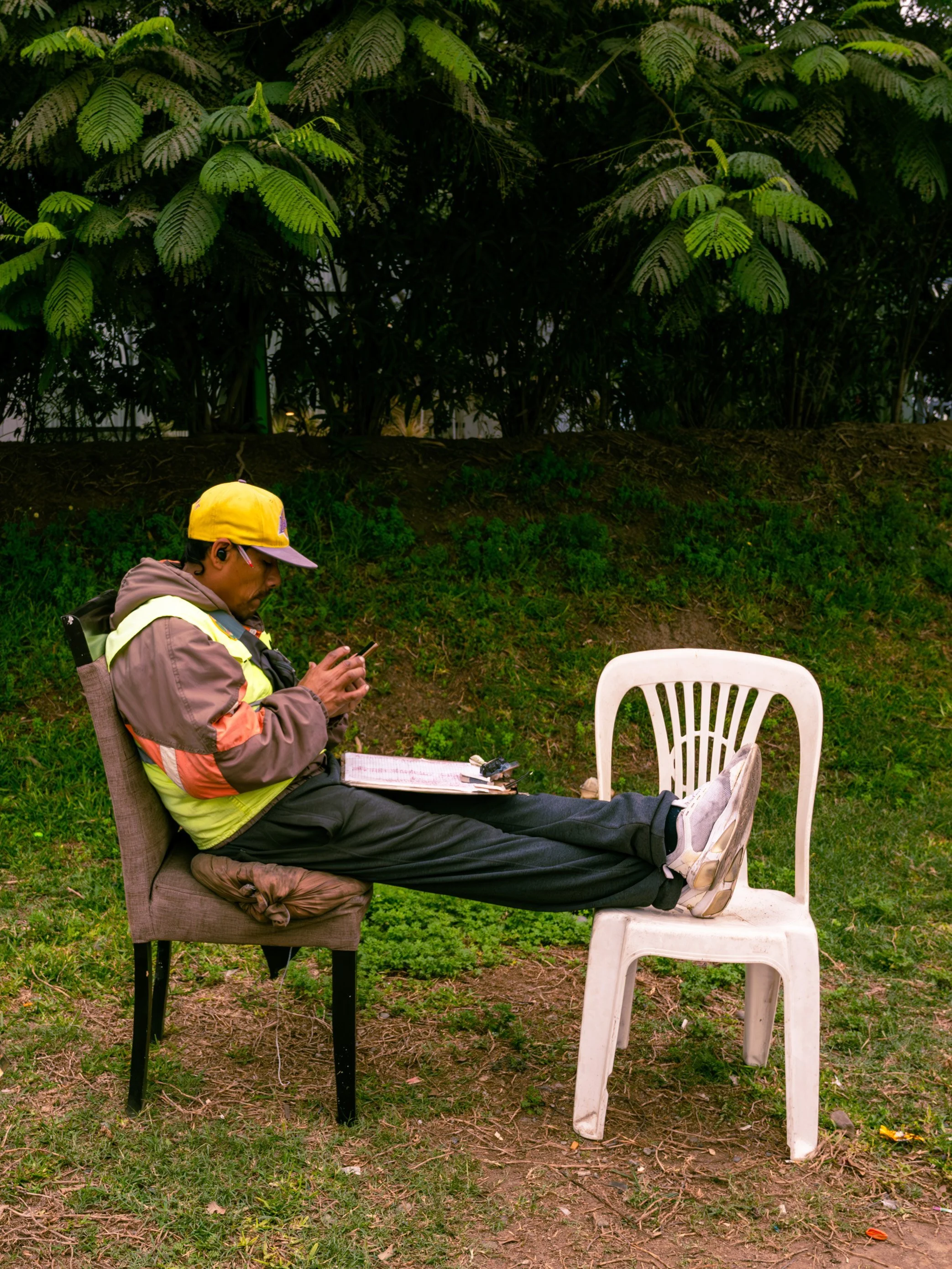 A man sitting in a chair with his feet resting on a plastic chair, playing on a phone, outdoors with greenery and trees in the background.