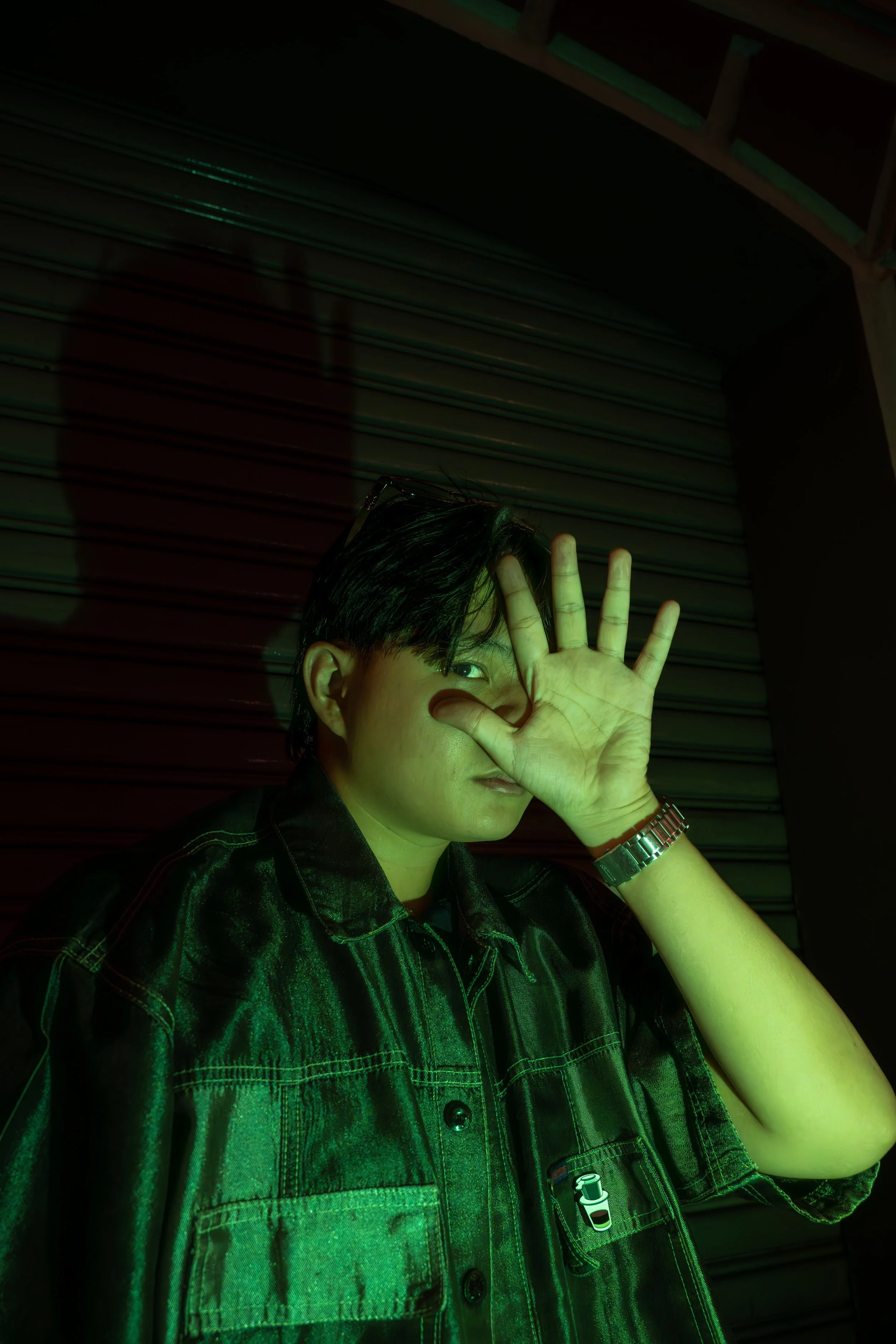 A person with black hair and glasses, wearing a checkered shirt and a wristwatch, is raising their hand in front of their face, partially covering one eye. The person is standing in front of a metal shutter door and is illuminated with green light.