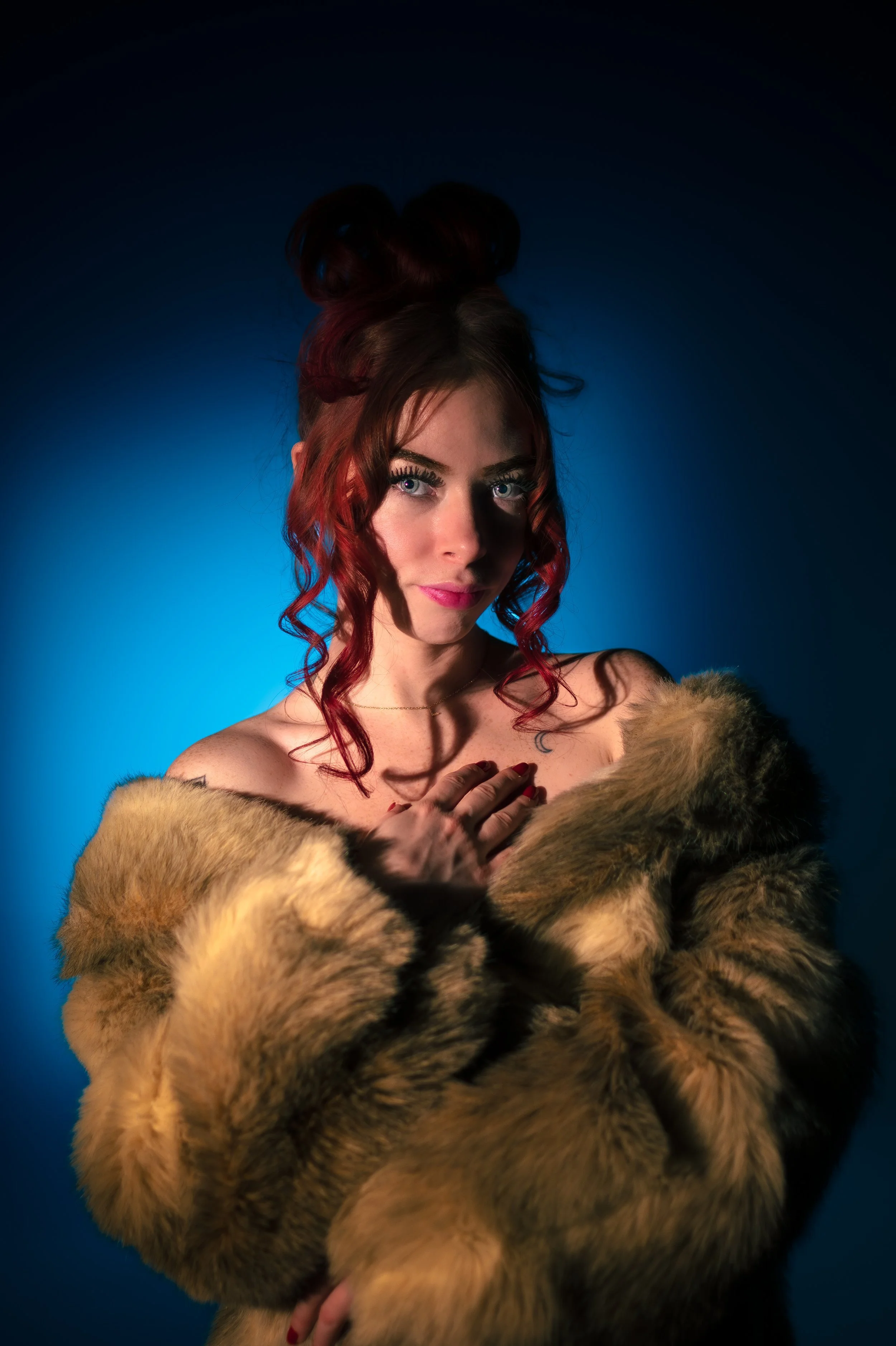 A woman with red hair styled in large buns, wearing a fur coat and makeup, posing against a blue background.
