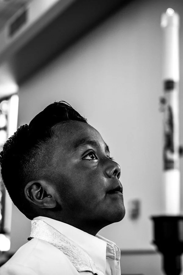 A young boy with short, styled hair looking upward inside a classroom or hall, with a large candles in the background.