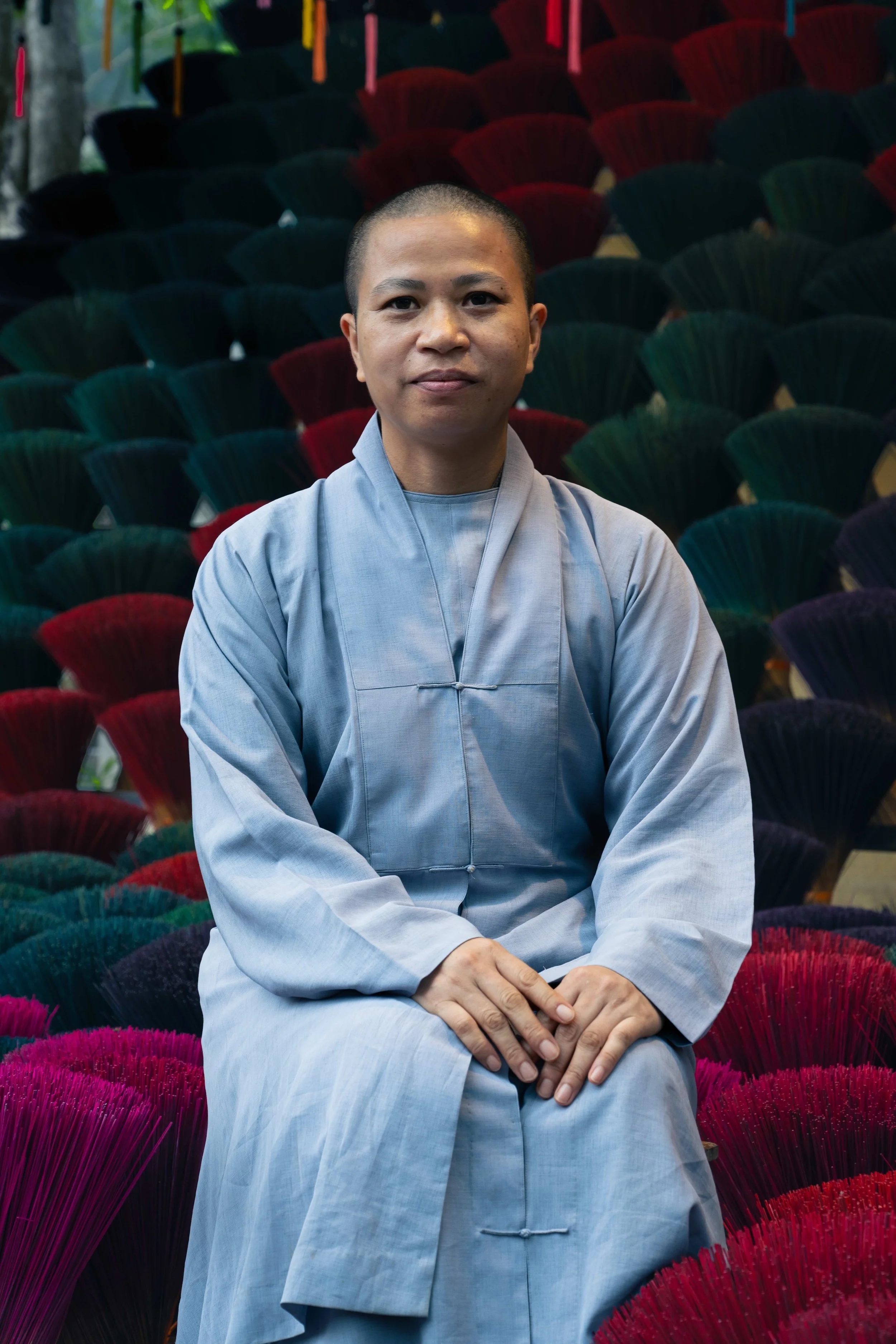 A person dressed in traditional Buddhist monk attire sitting with hands folded in lap, surrounded by colorful fanned umbrellas or decorations in shades of green, red, purple, and pink.