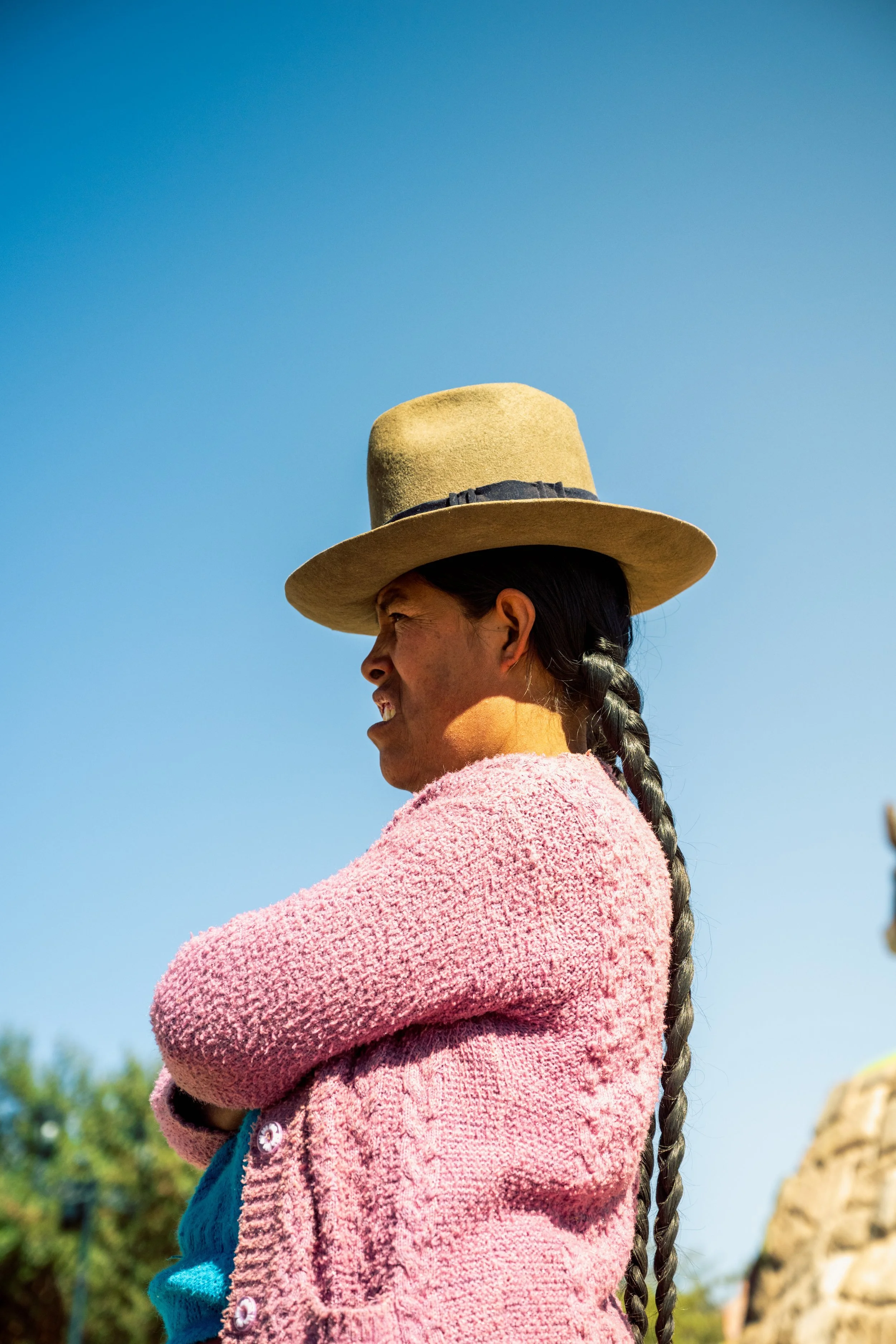 Profile of a woman with braided hair wearing a wide-brimmed hat and a pink fuzzy sweater, standing outdoors against a clear blue sky.
