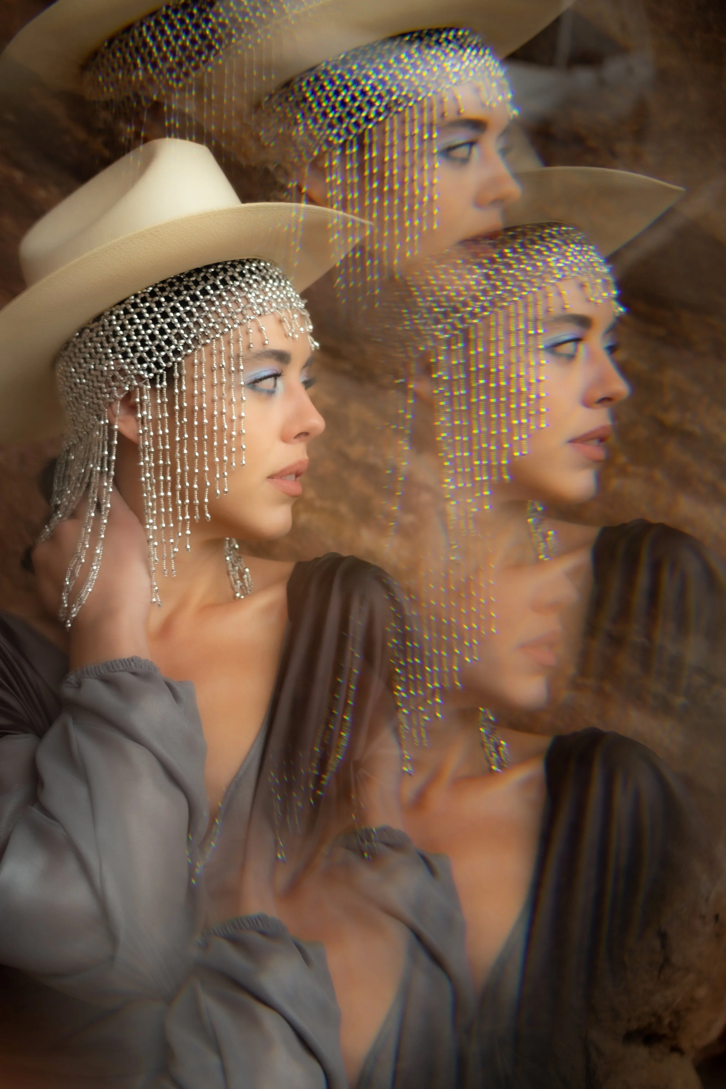 A woman with blonde hair wearing a large pearl and rhinestone headpiece, beige wide-brimmed hat, and black top with sheer sleeves, posing against a textured brown background. The image has a glitch effect with rainbow-colored pixelation.
