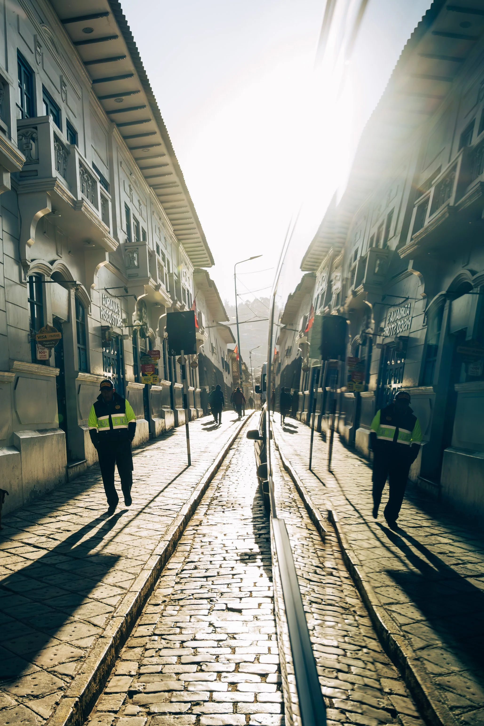 A narrow cobblestone street with tram tracks, flanked by historic white buildings with decorative balconies and blue balconies, under a bright sky with sunlight glare, and several people walking along the sidewalk.