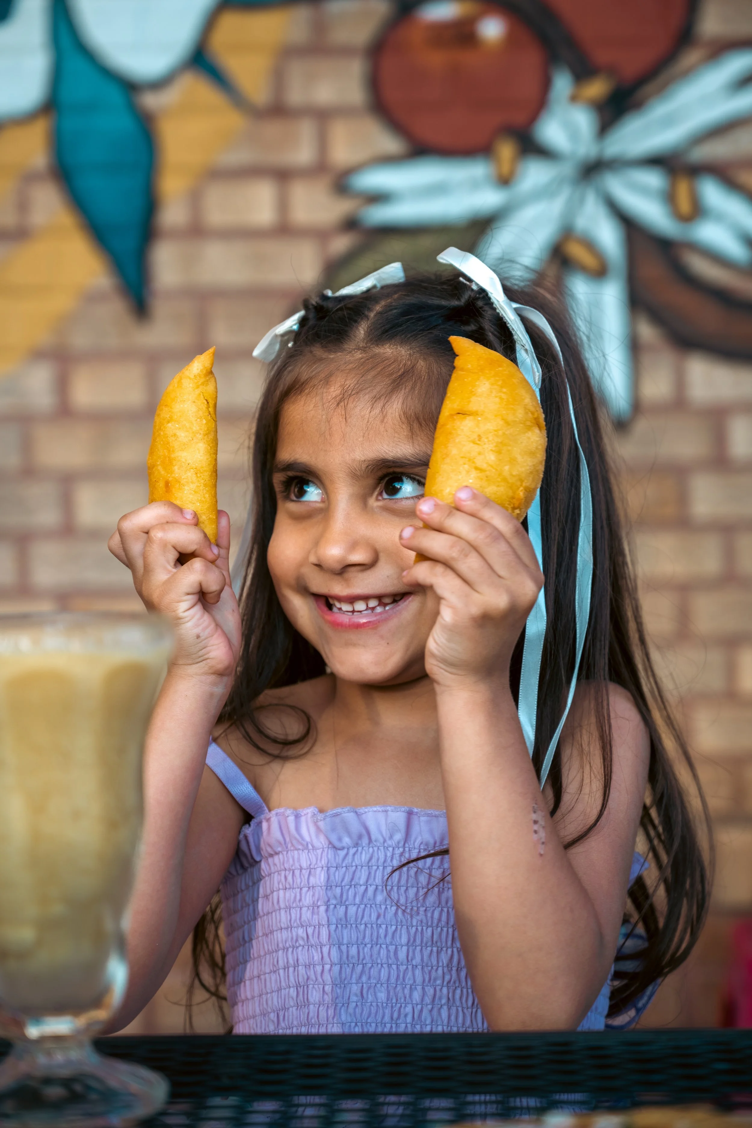 A young girl with long dark hair wearing a light purple dress and a white bow in her hair, holding two potato chips up to her head like ears, smiling, with a colorful butterfly mural on a brick wall in the background.