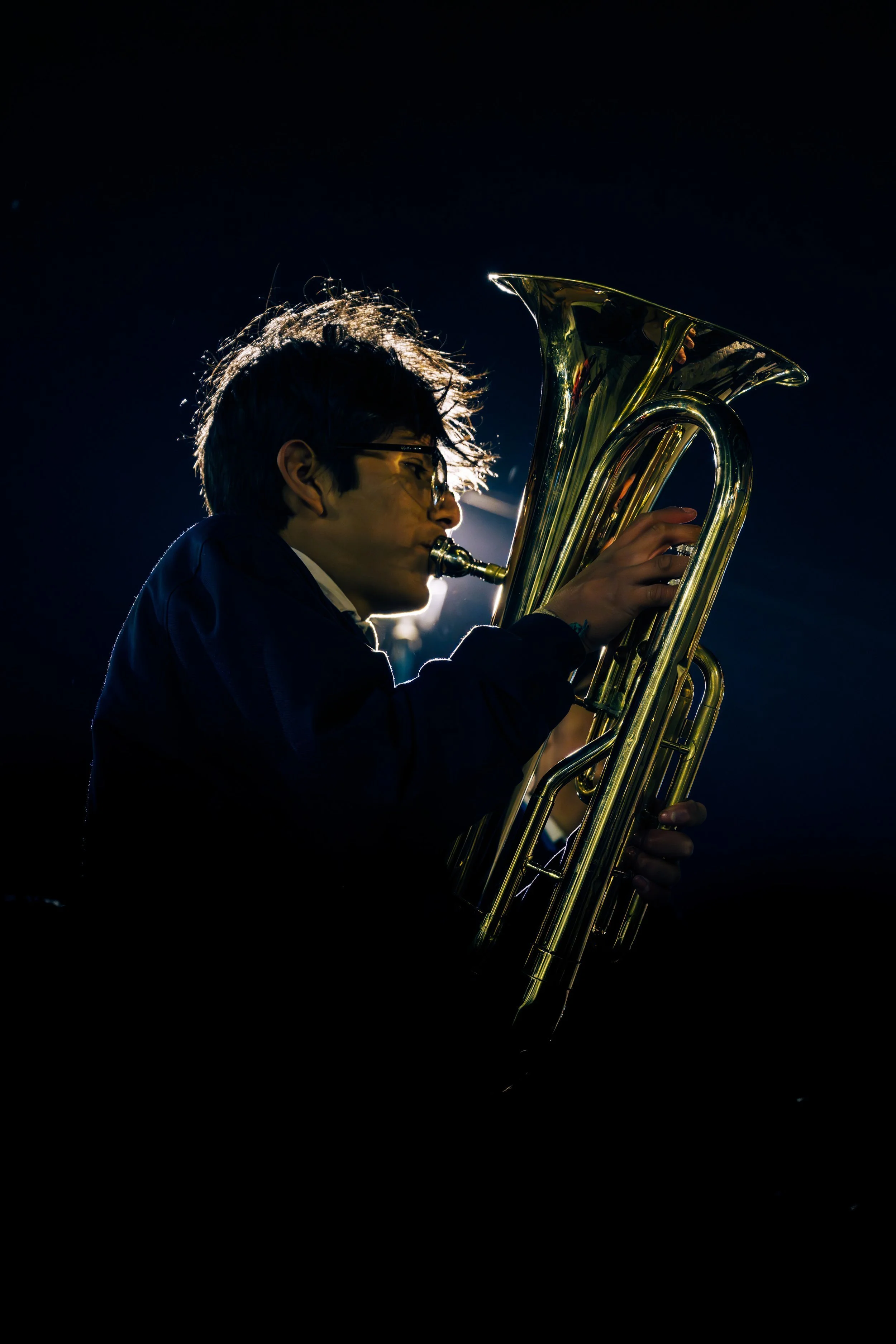 A person playing a shiny brass euphonium, facing to the left, under dramatic lighting with a dark background.