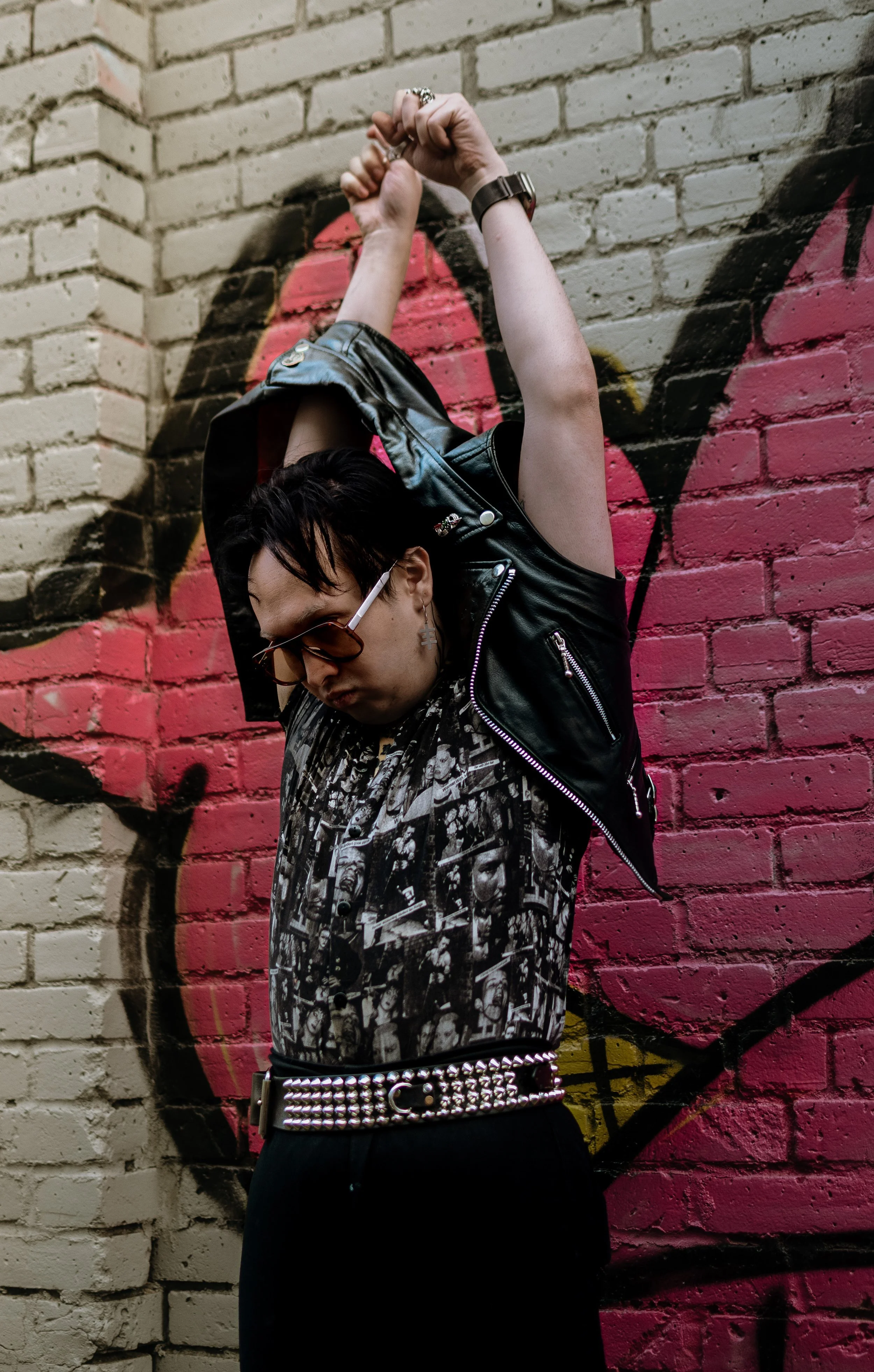 Person with short dark hair wearing sunglasses, a black leather vest with metal zippers, a printed shirt, a studded belt, and multiple rings, standing in front of a brick wall with pink graffiti art.
