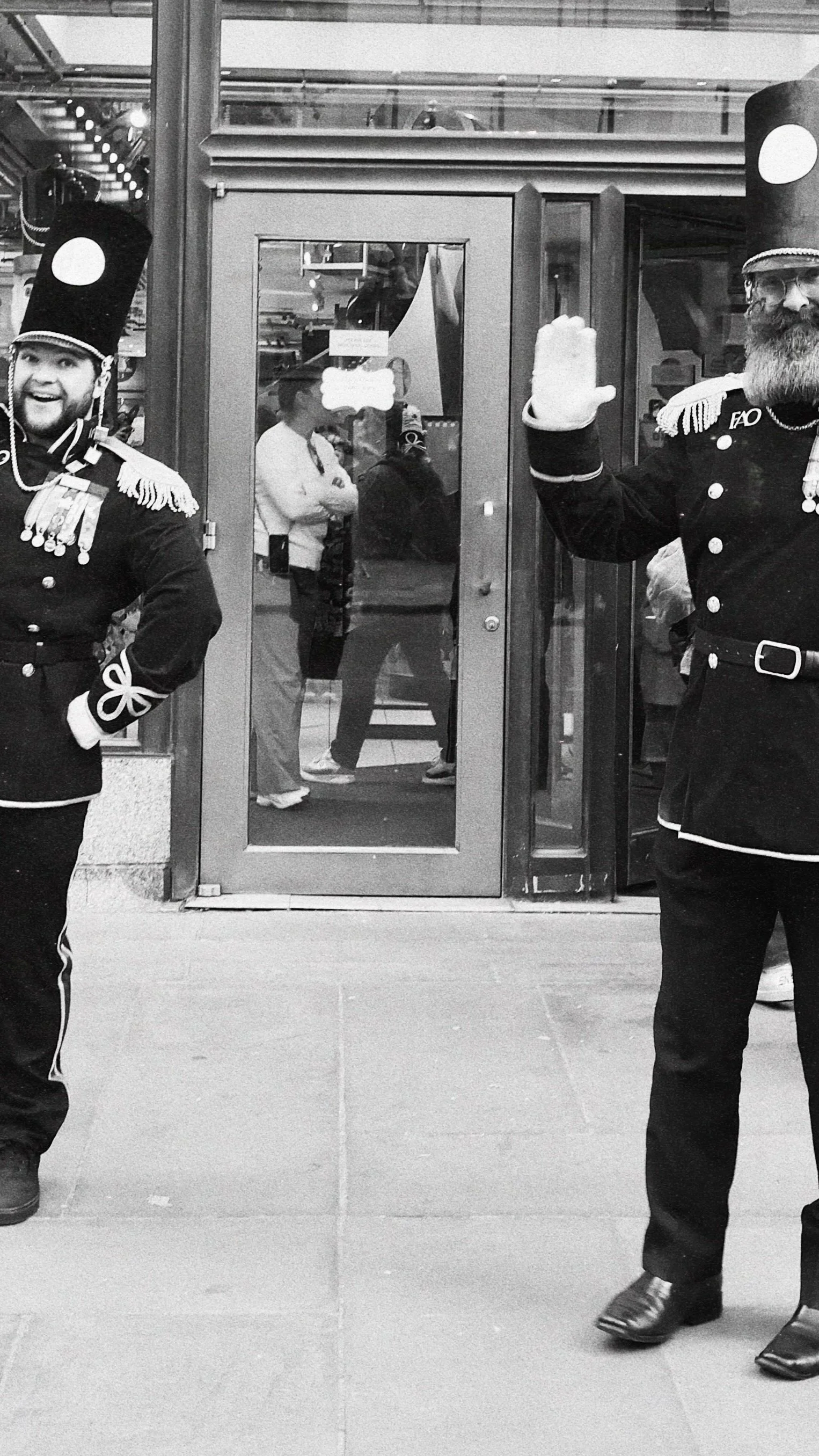 Two men dressed as toy soldiers or drummer boys standing outside a shop entrance, one smiling and waving.