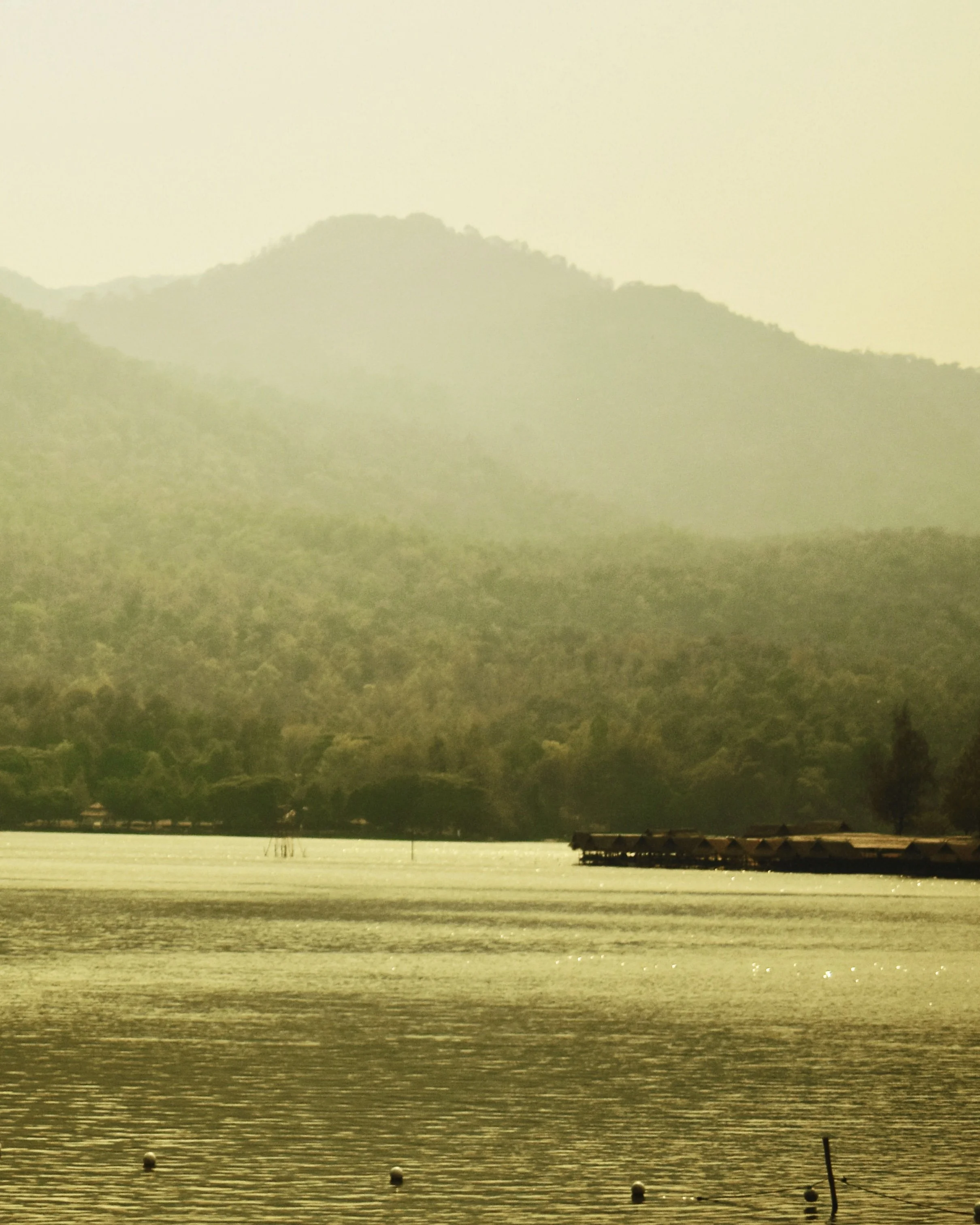 A serene landscape with a lake, forested hills, and mountains in the background, under a yellowish sky.
