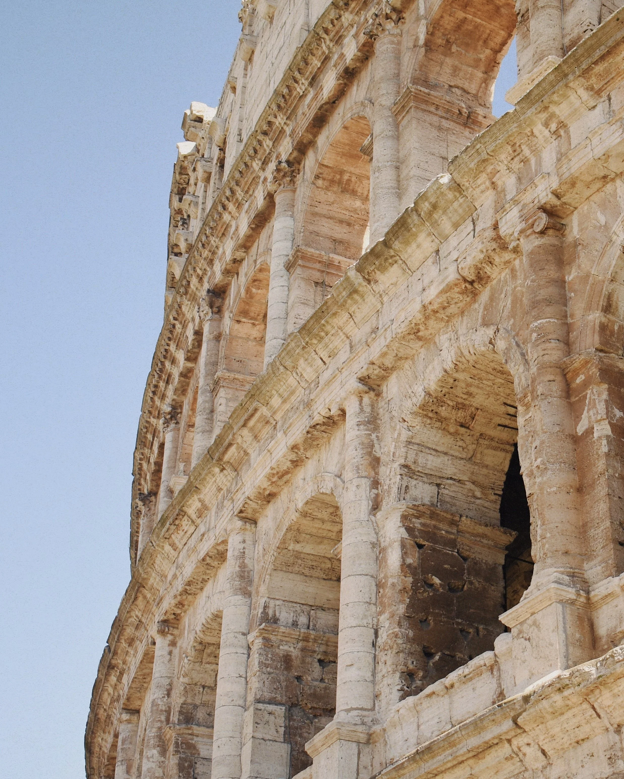 Close-up of the ancient stone exterior of the Roman Colosseum showing arches and columns against a clear blue sky.