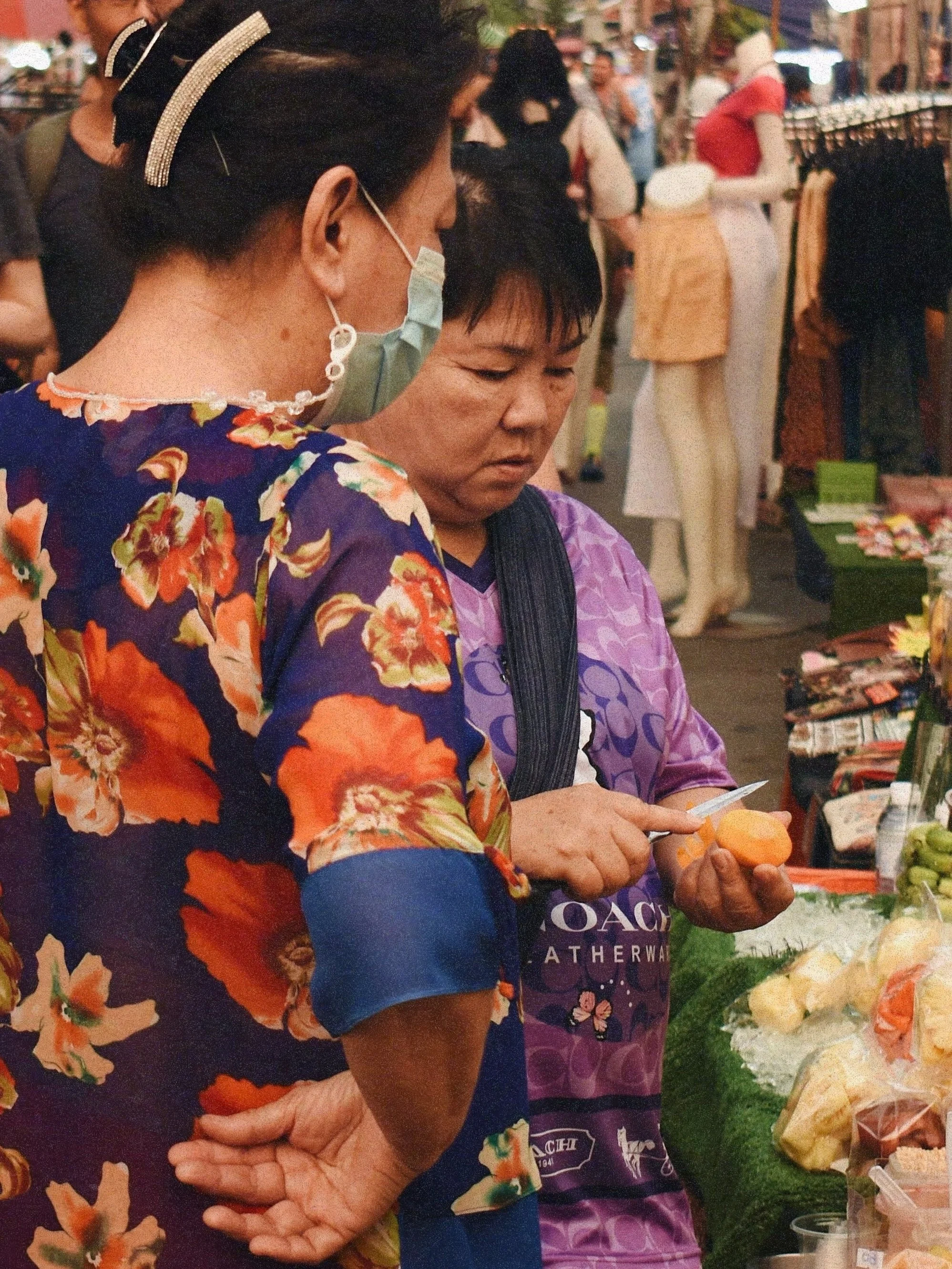 Two women shopping at an outdoor market, one woman in a floral dress with a face mask, and the other in a purple apron, holding a piece of fruit and examining it.