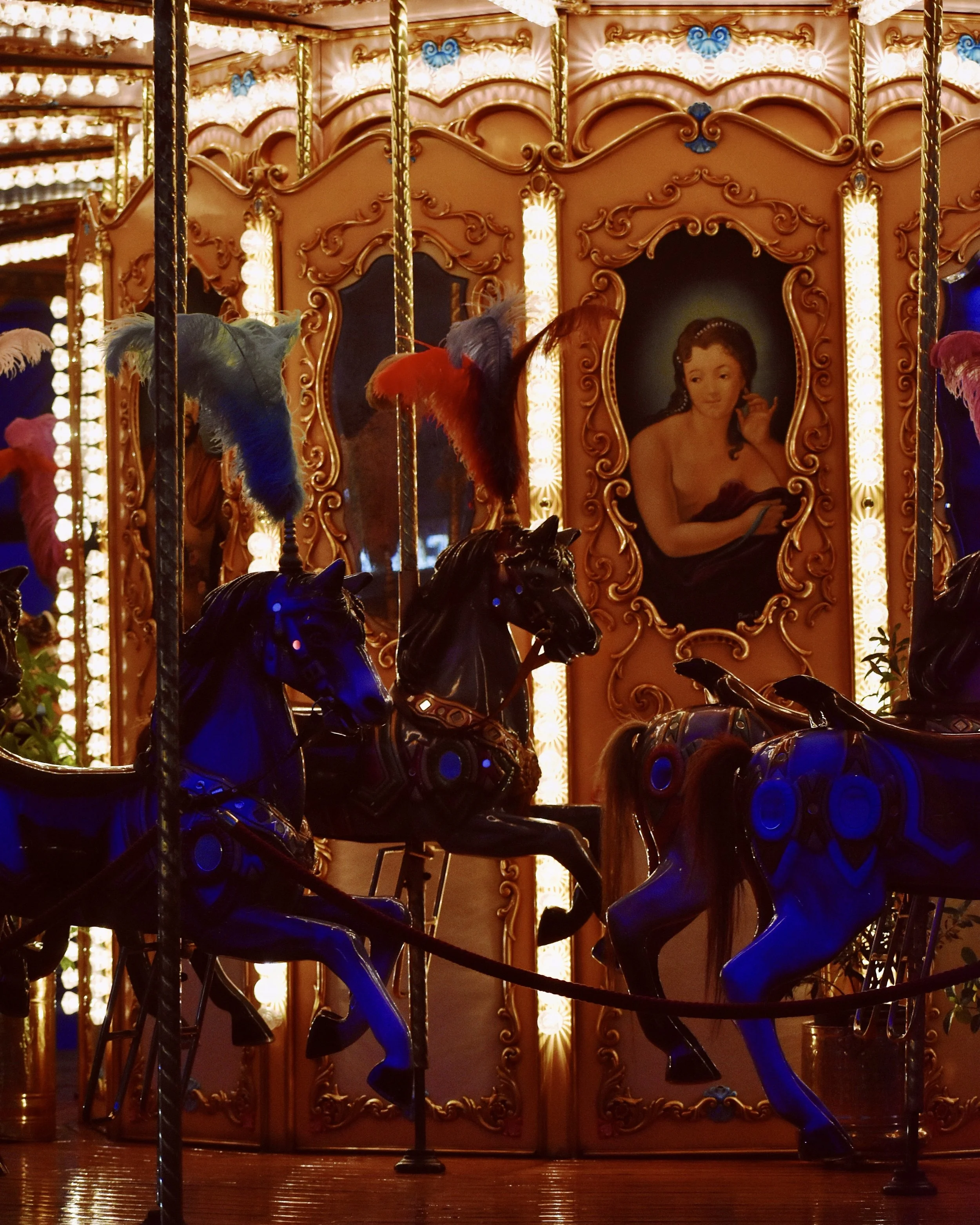 A vintage carousel with painted horses and a woman painted in a portrait in the background, illuminated by warm lights.
