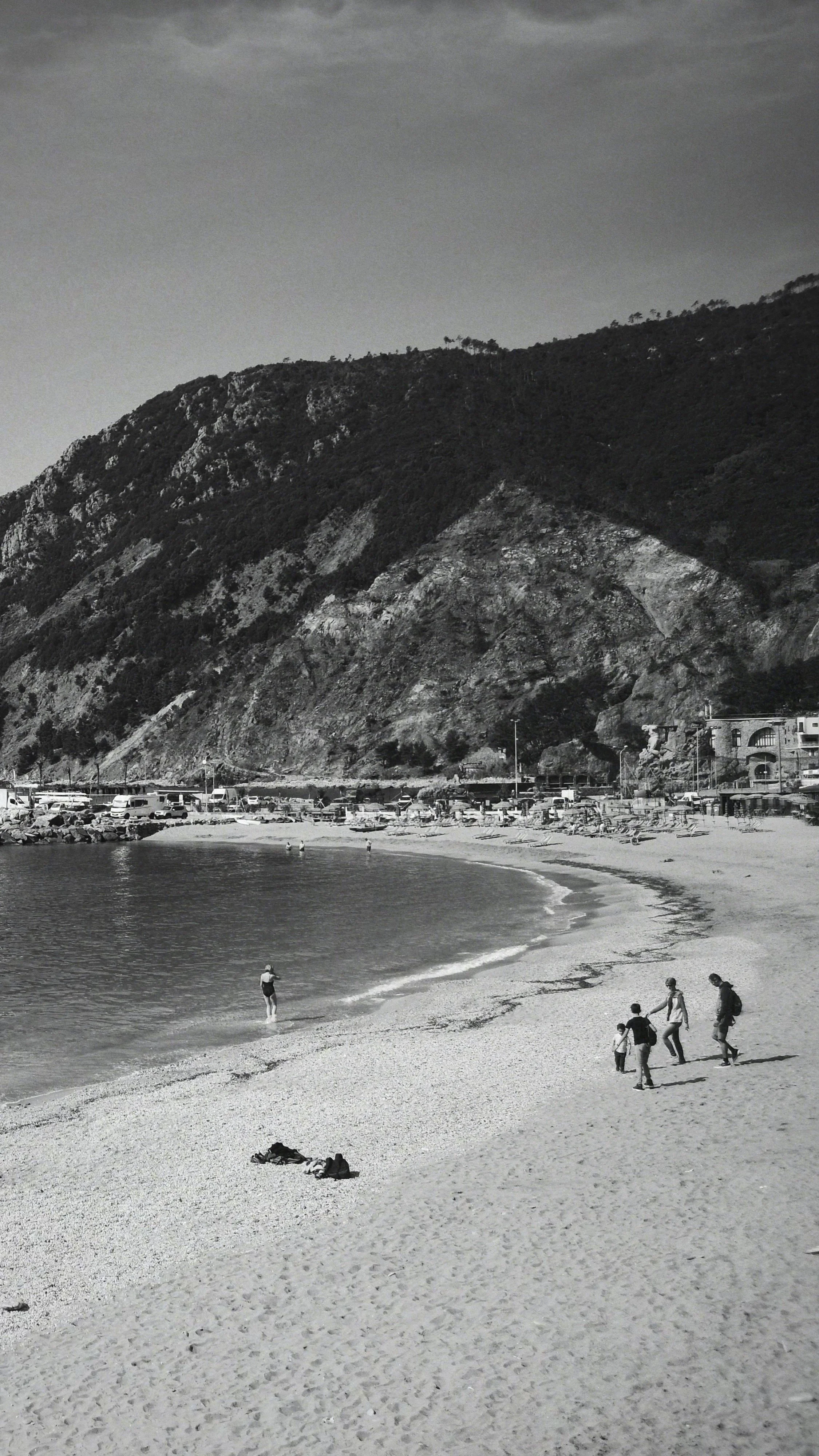 A black and white photo of a beach with four people walking along the shoreline and one person swimming in the water. There are mountains in the background with some buildings and parked cars at the base.