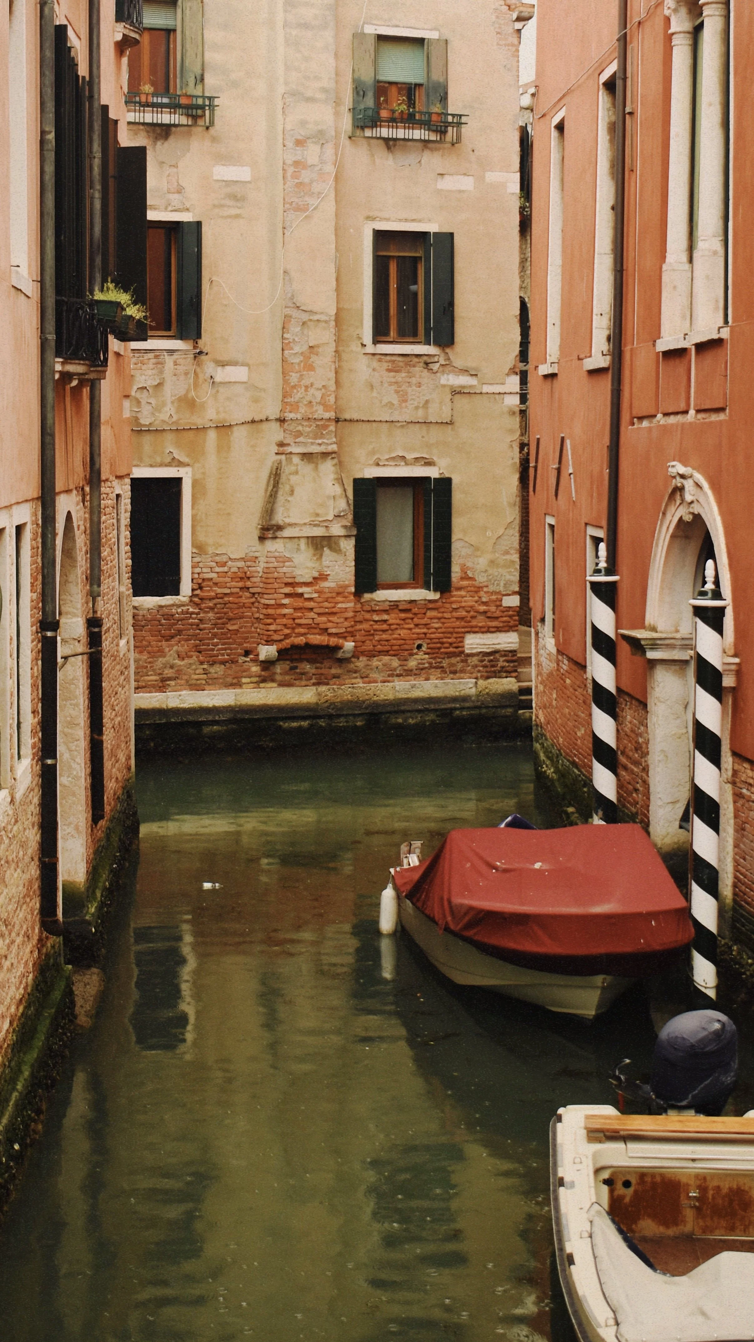 A narrow canal with two boats, one with a red cover, between old brick buildings with windows and green shutters in Venice, Italy.