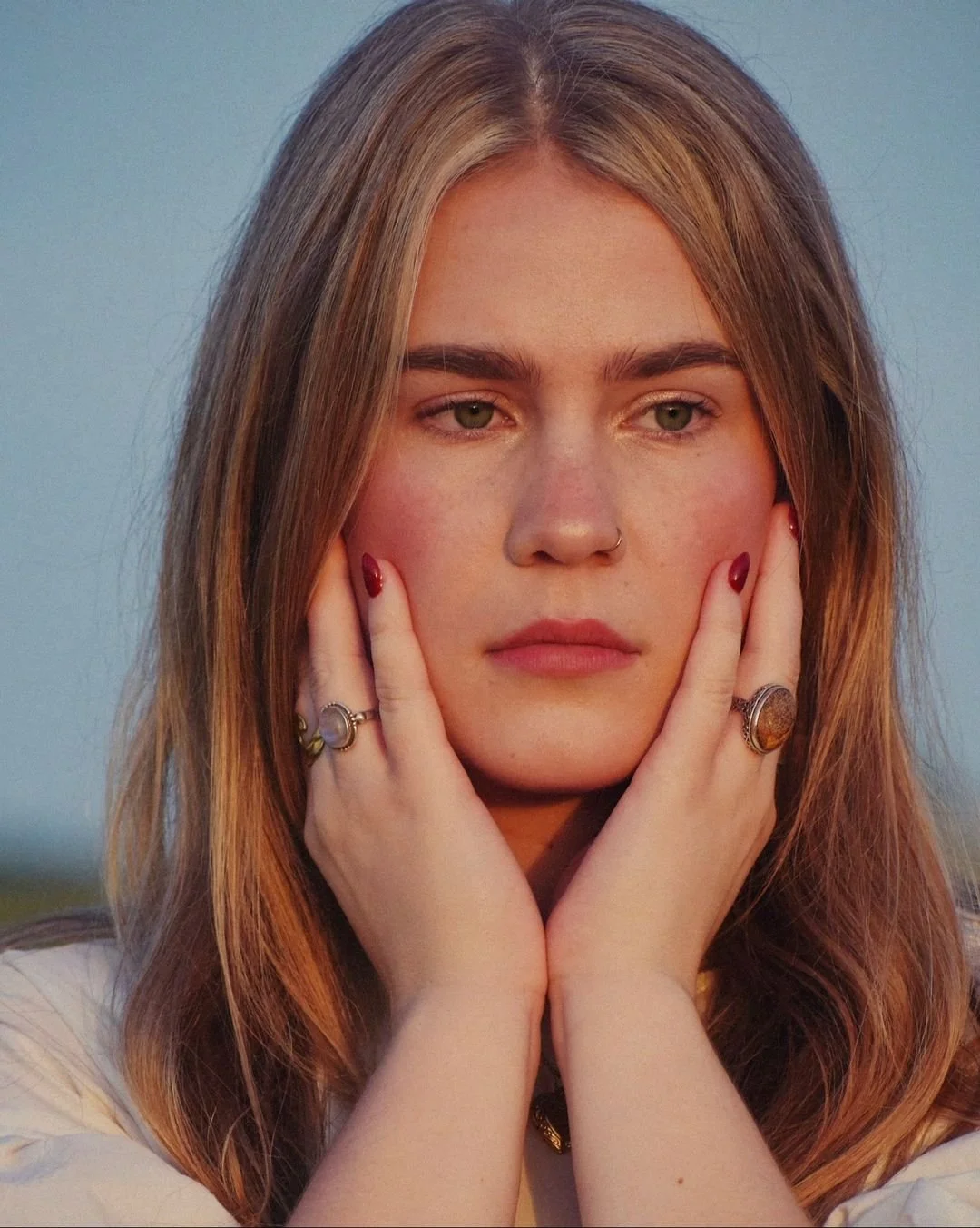 A young woman with long, light brown hair and green eyes, holding her face with both hands, wearing rings and a nose ring, looking contemplative.