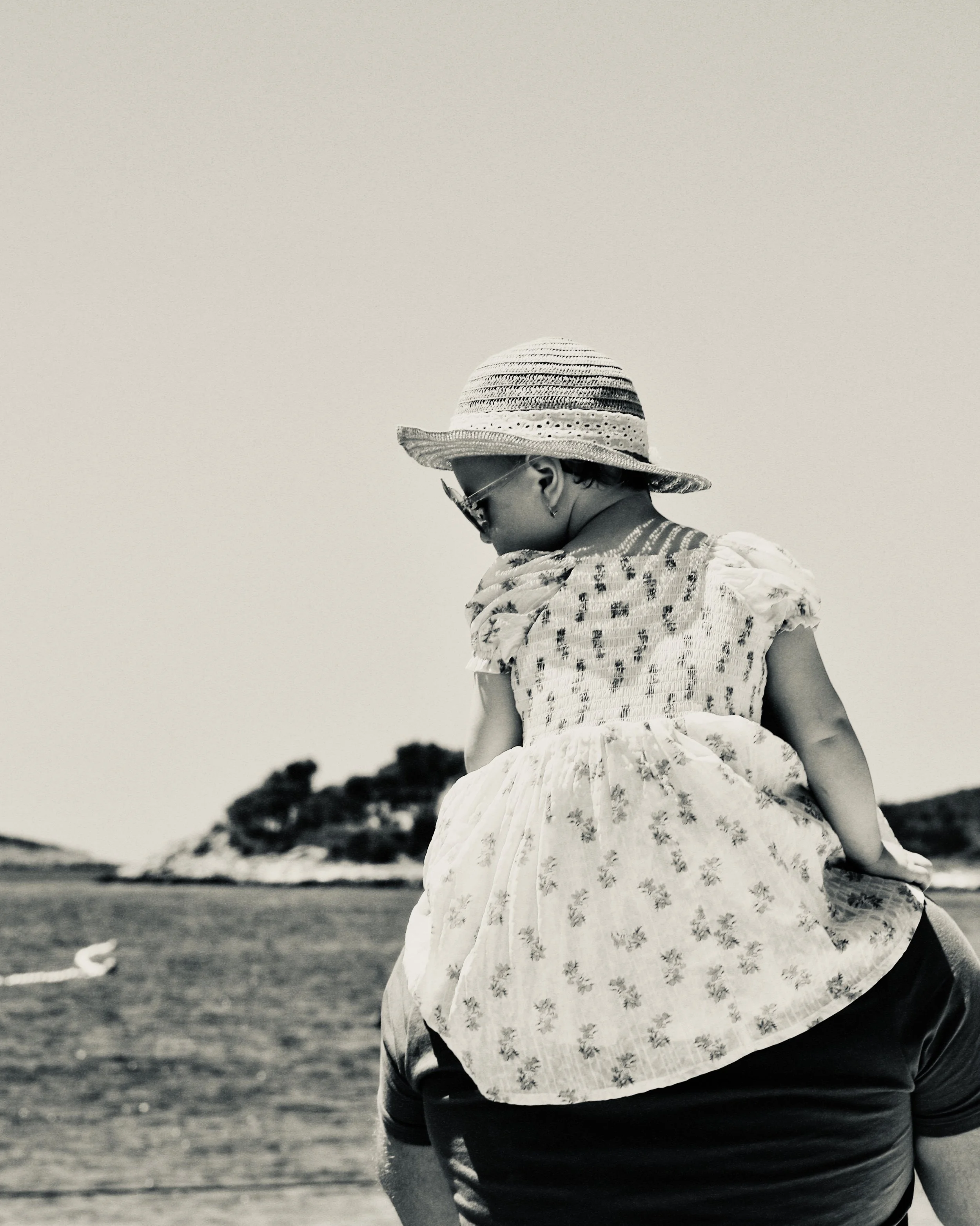 Child wearing a hat and sunglasses being carried on someone's shoulders at the beach, with ocean waves and a small island in the background.