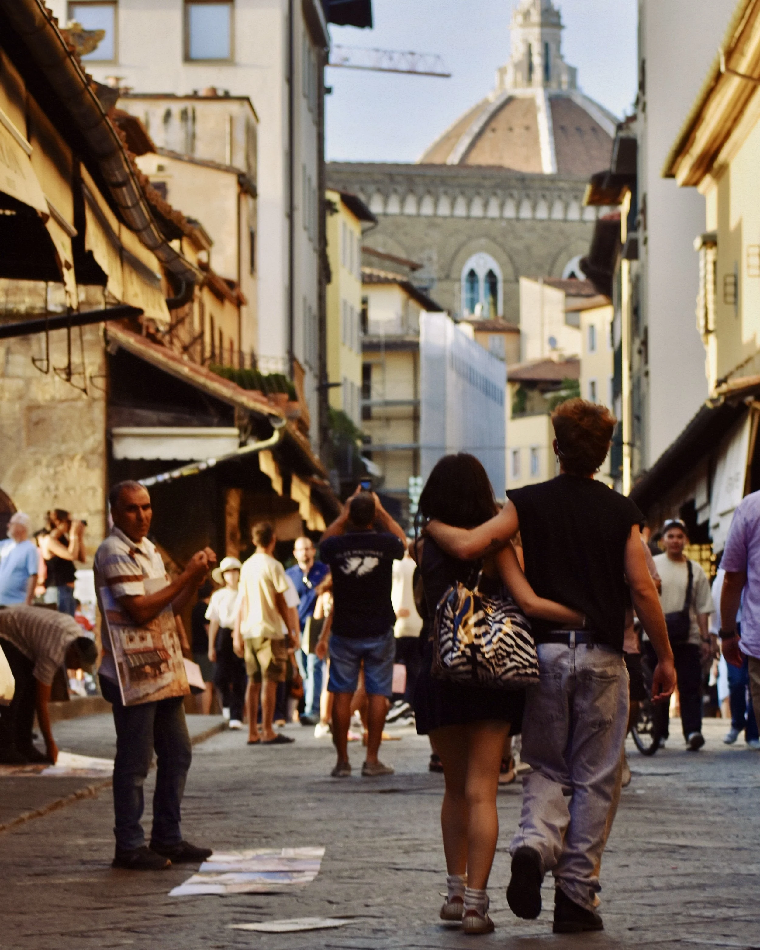A crowded street scene with people walking and taking photos, old buildings with awnings, and a large domed church in the background.