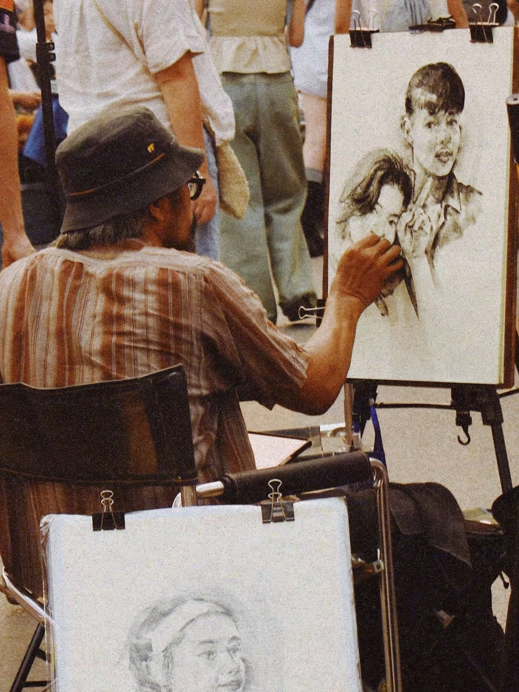 An elderly male artist wearing a striped shirt and a wide-brimmed hat, seated with his back to the camera, drawing a portrait of a young girl and a boy on a large piece of paper. The artist is using a pencil or charcoal, and there are in-progress sketches on other sheets of paper nearby. The scene is in a busy art event or exhibit with people standing around.