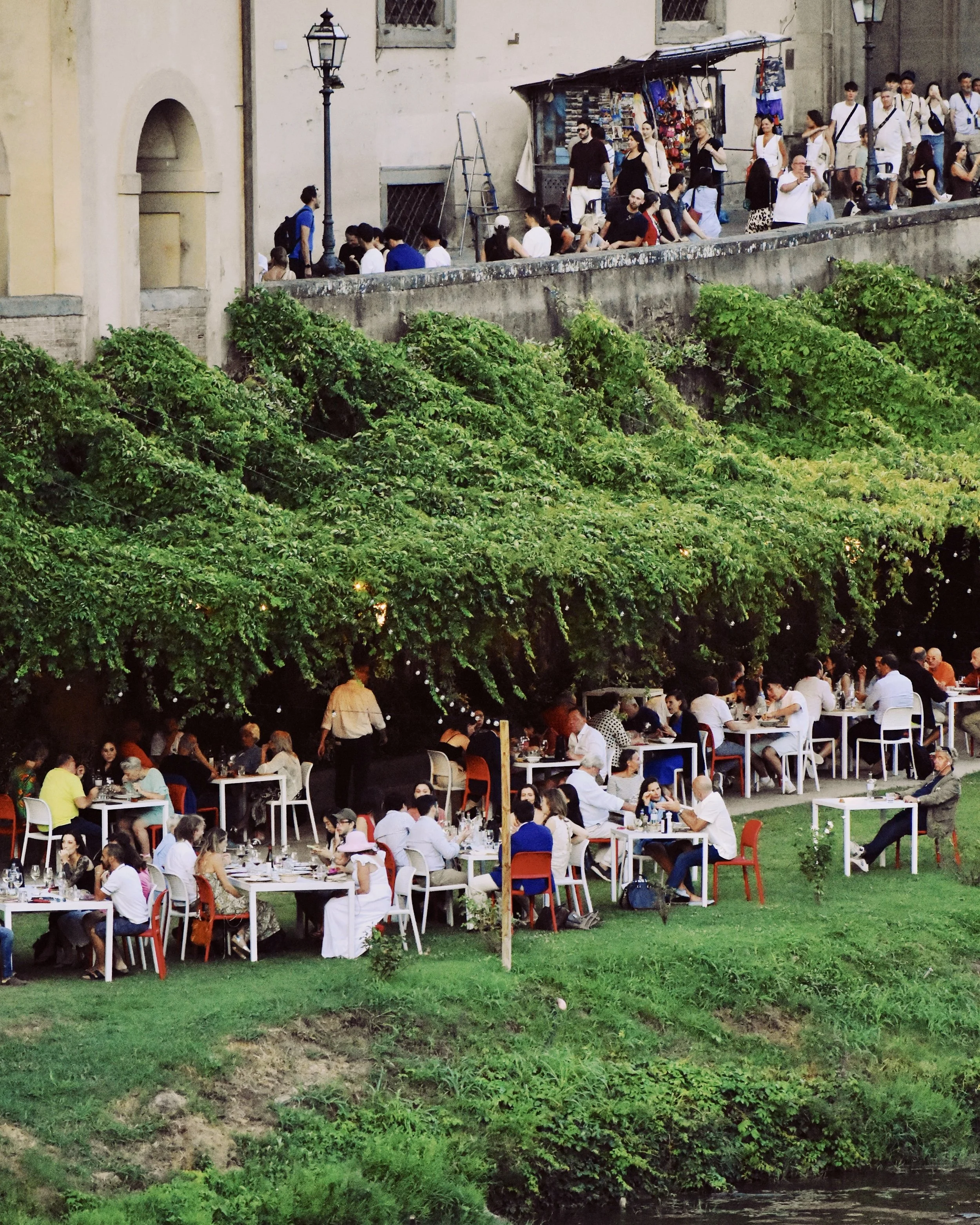Outdoor dining area with tables and chairs filled with people, set under green treetops and adjacent to a grassy area and a river.