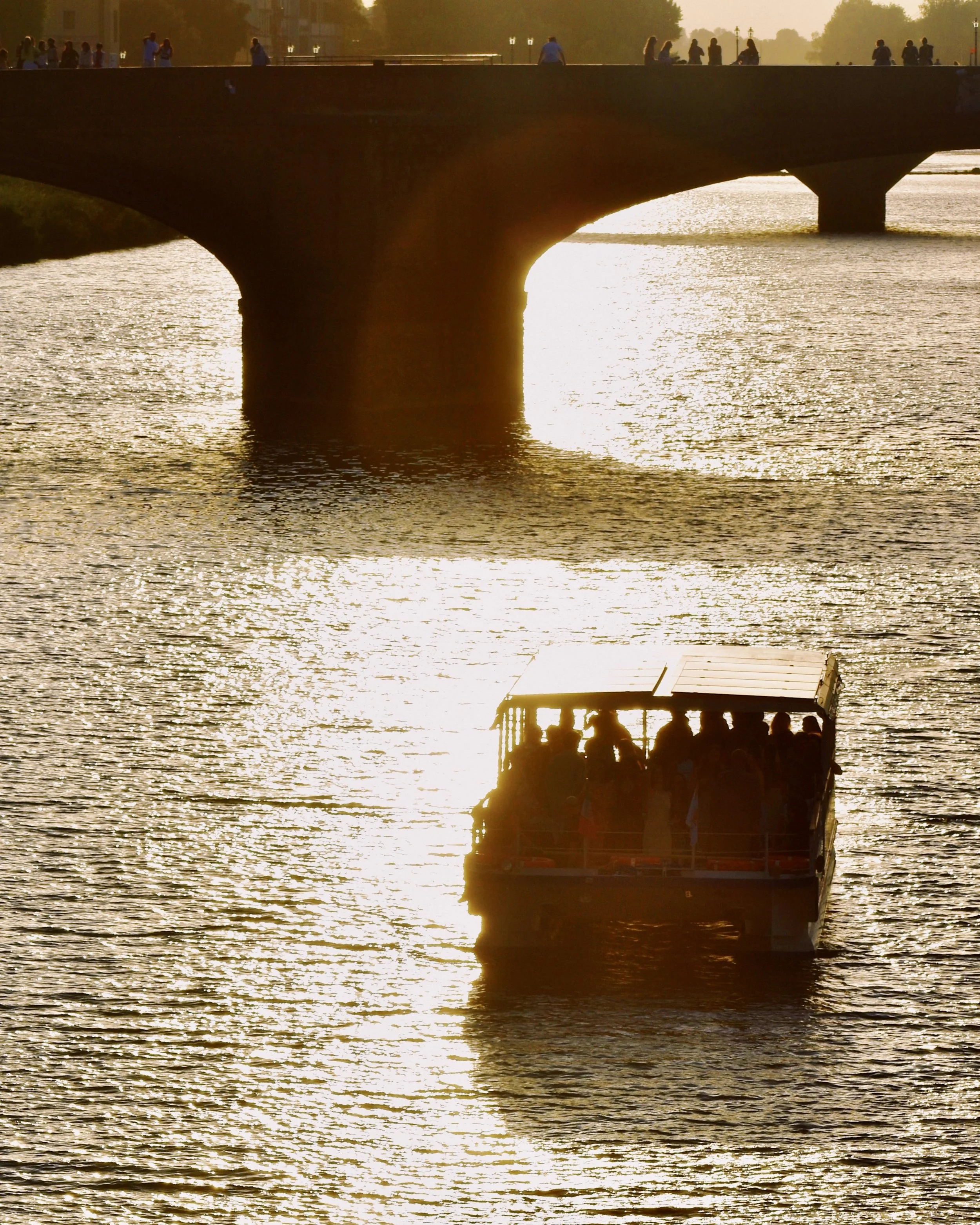 A boat with people on board sailing on a river at sunset, with a bridge and people on it in the background