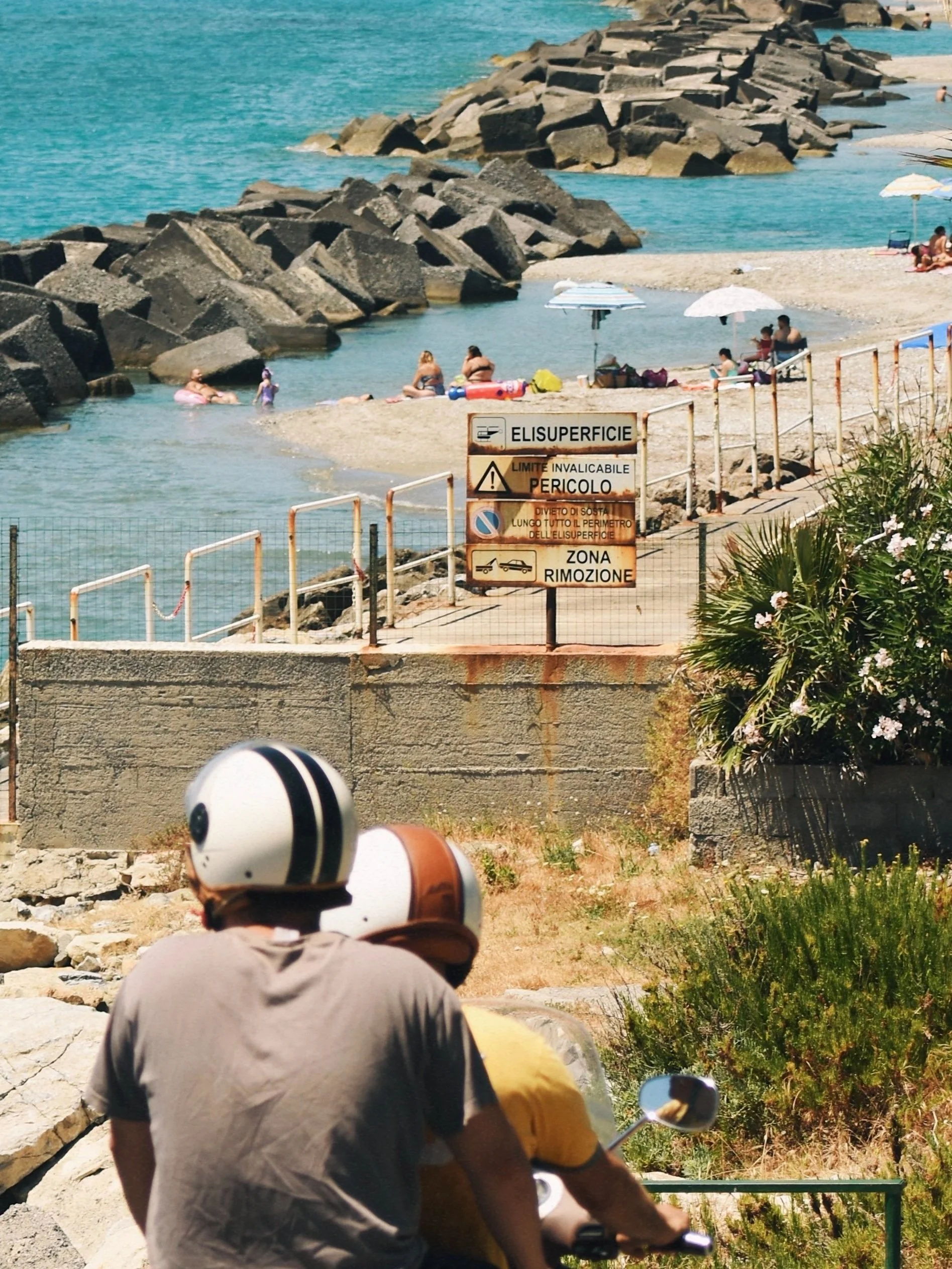 View of a small beach with people swimming, lounging under umbrellas, and rocky breakwaters. A sign in Italian indicates the surface area and removal zone, with a couple of motorcyclists in the foreground.