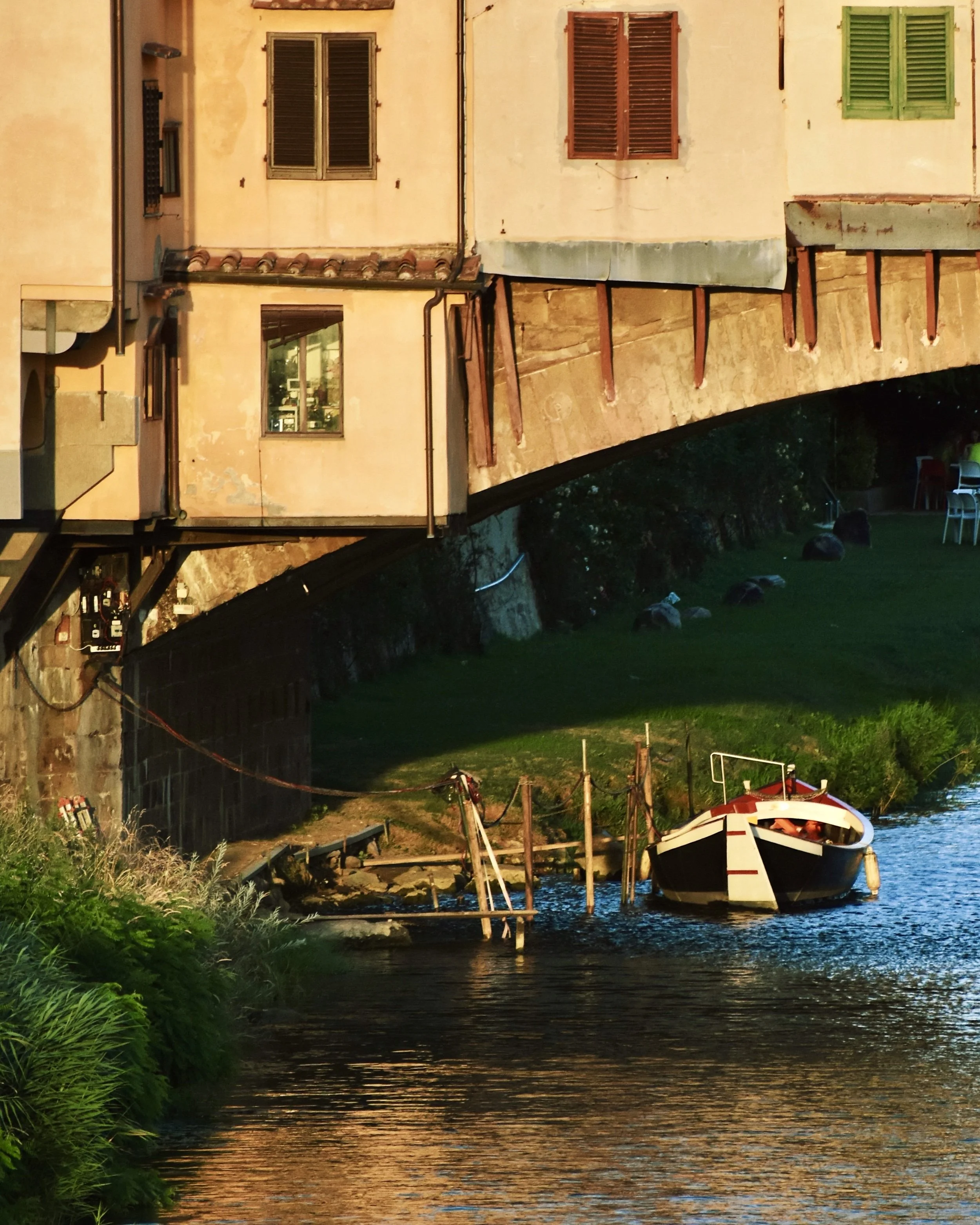 A house built on a bridge over a river with a dock and a small boat tied to it, surrounded by greenery.