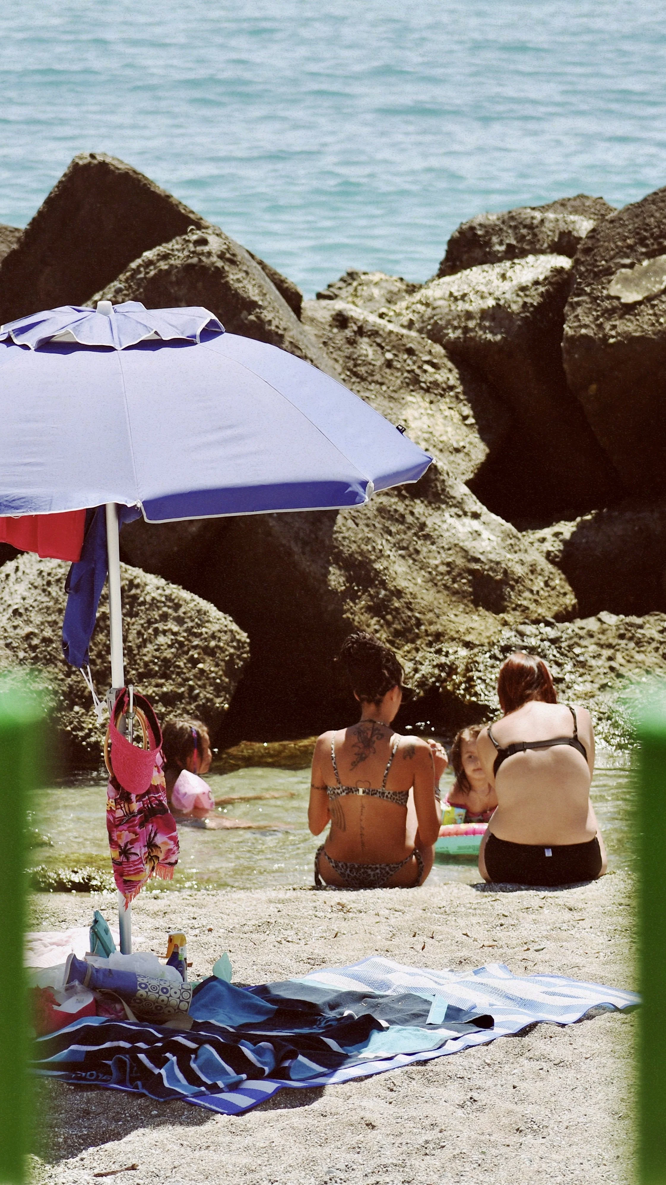 People relaxing on a beach with rocks and water in the background, under a blue umbrella.