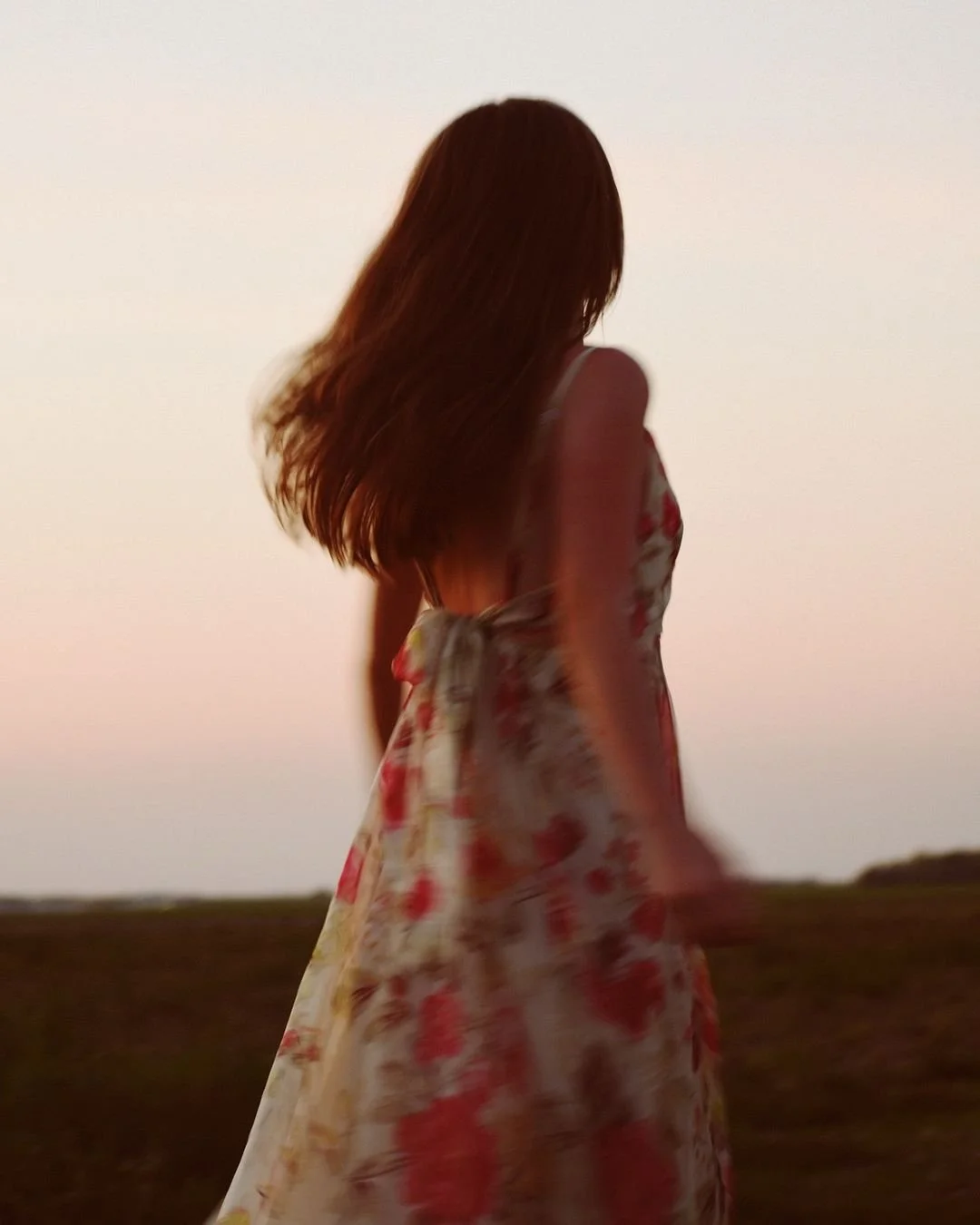 Silhouette of a woman with long hair standing outdoors at sunset, wearing a floral dress.