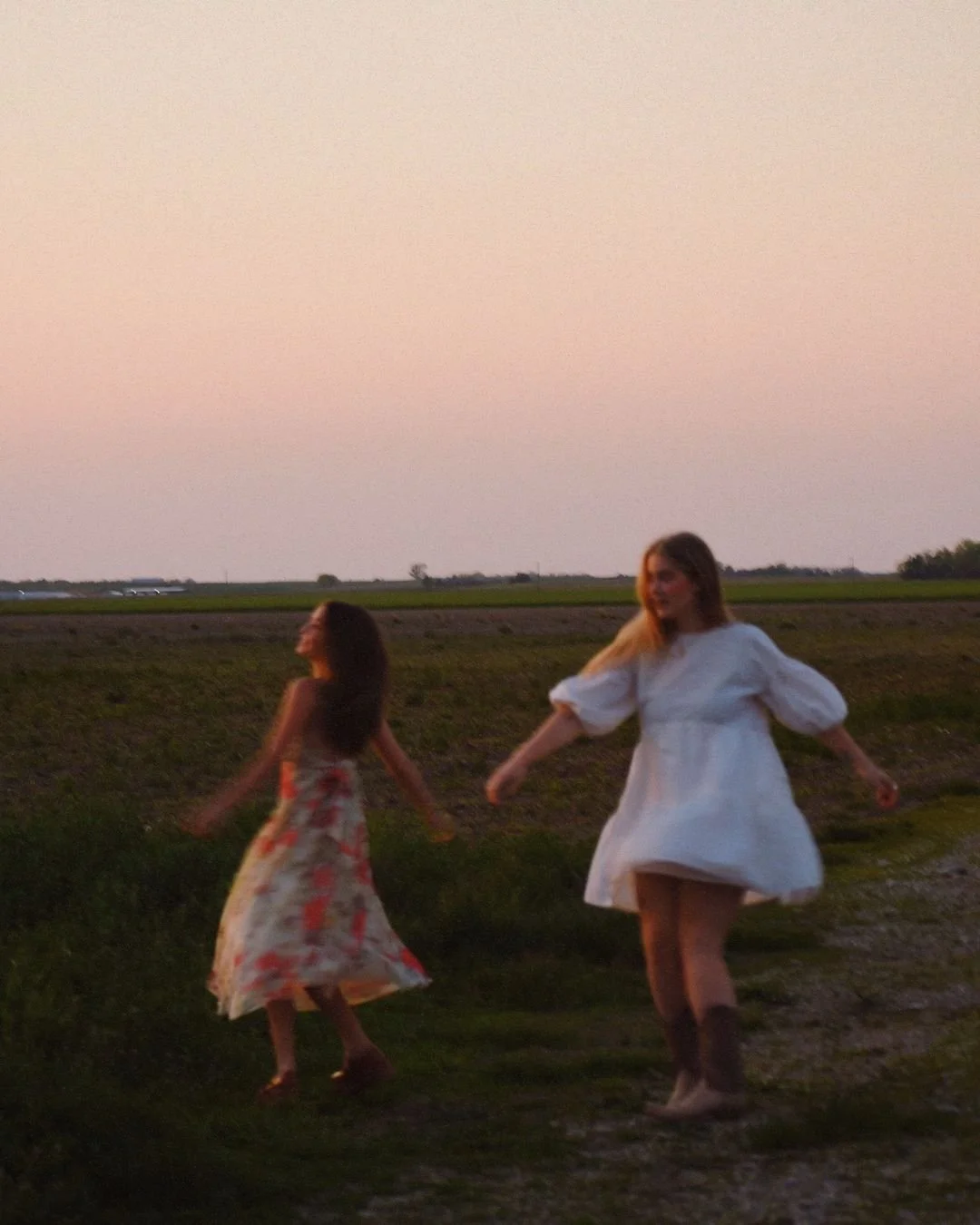 Two women dancing or twirling outdoors during sunset, wearing dresses, one in a floral gown and the other in a white dress with puffy sleeves, in a grassy rural area.