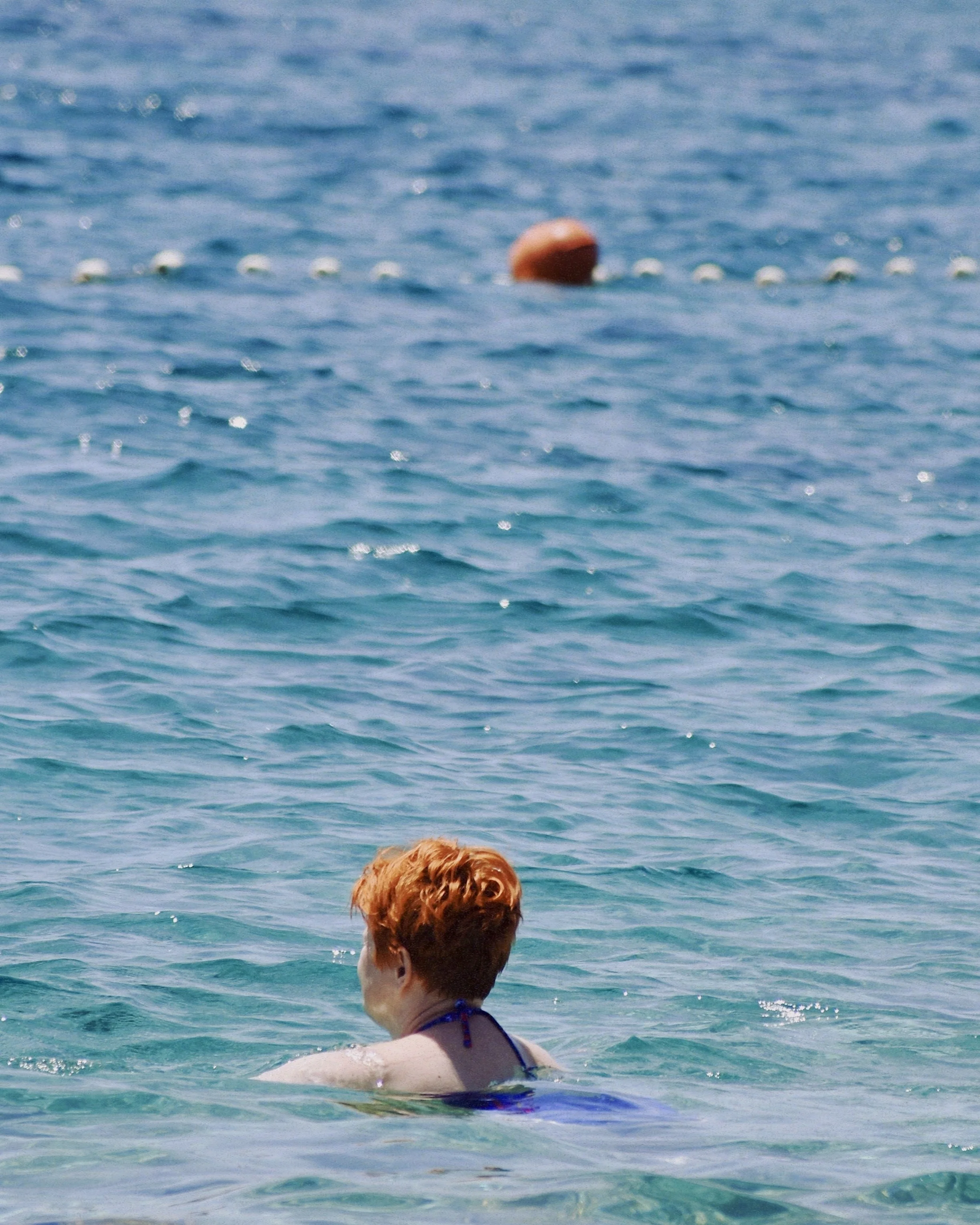 Person with red hair swimming in the ocean facing away, with a buoy in the water nearby.