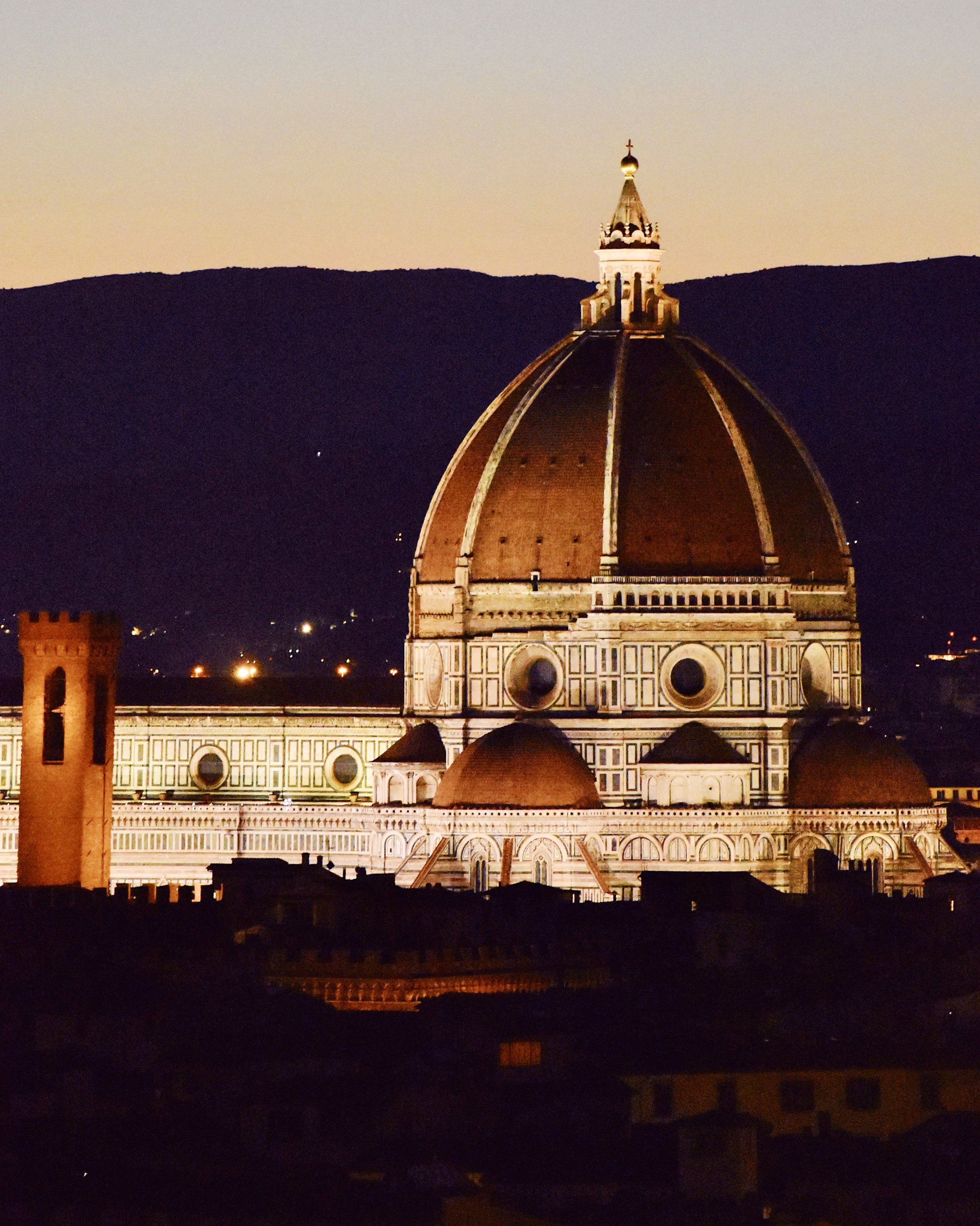 Nighttime view of Florence Cathedral with its large dome illuminated against a dark sky and mountains in the background.