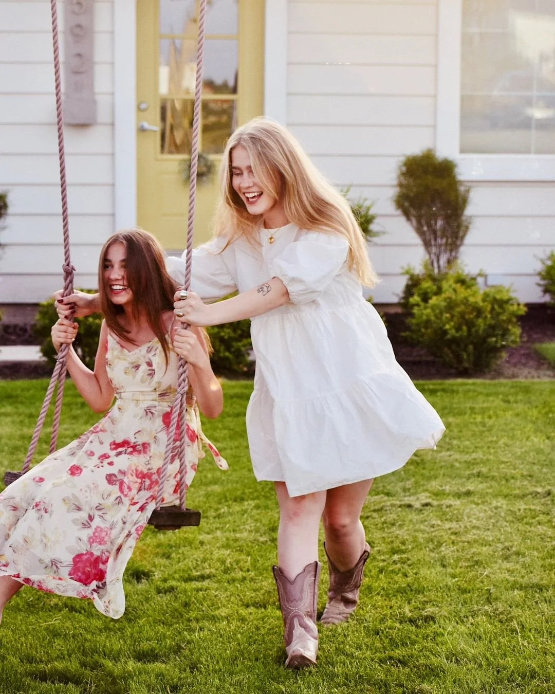 Two young women with long hair, one with blonde and one with brown, playing on a swing set in a backyard, smiling and enjoying the sunny day. The blonde is standing, holding the swing ropes, while the brown-haired woman is sitting on the swing. The background features a white house with a yellow door and green bushes.