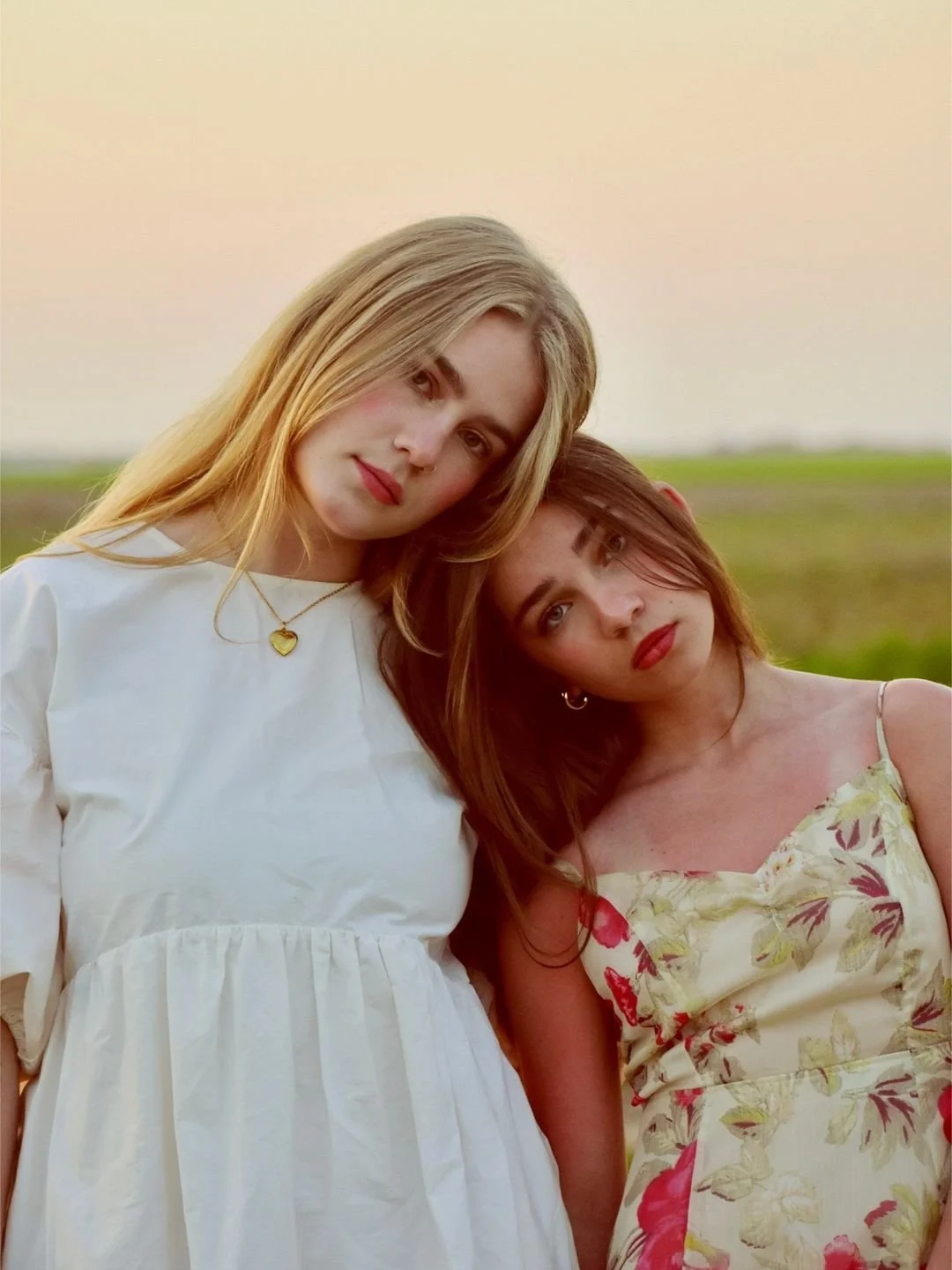 Two young women standing close together outdoors at sunset, with one resting her head on the other's shoulder. One woman has blonde hair and is wearing a white dress with a gold heart necklace, and the other has reddish-brown hair and is wearing a floral dress. They are looking at the camera with neutral expressions.