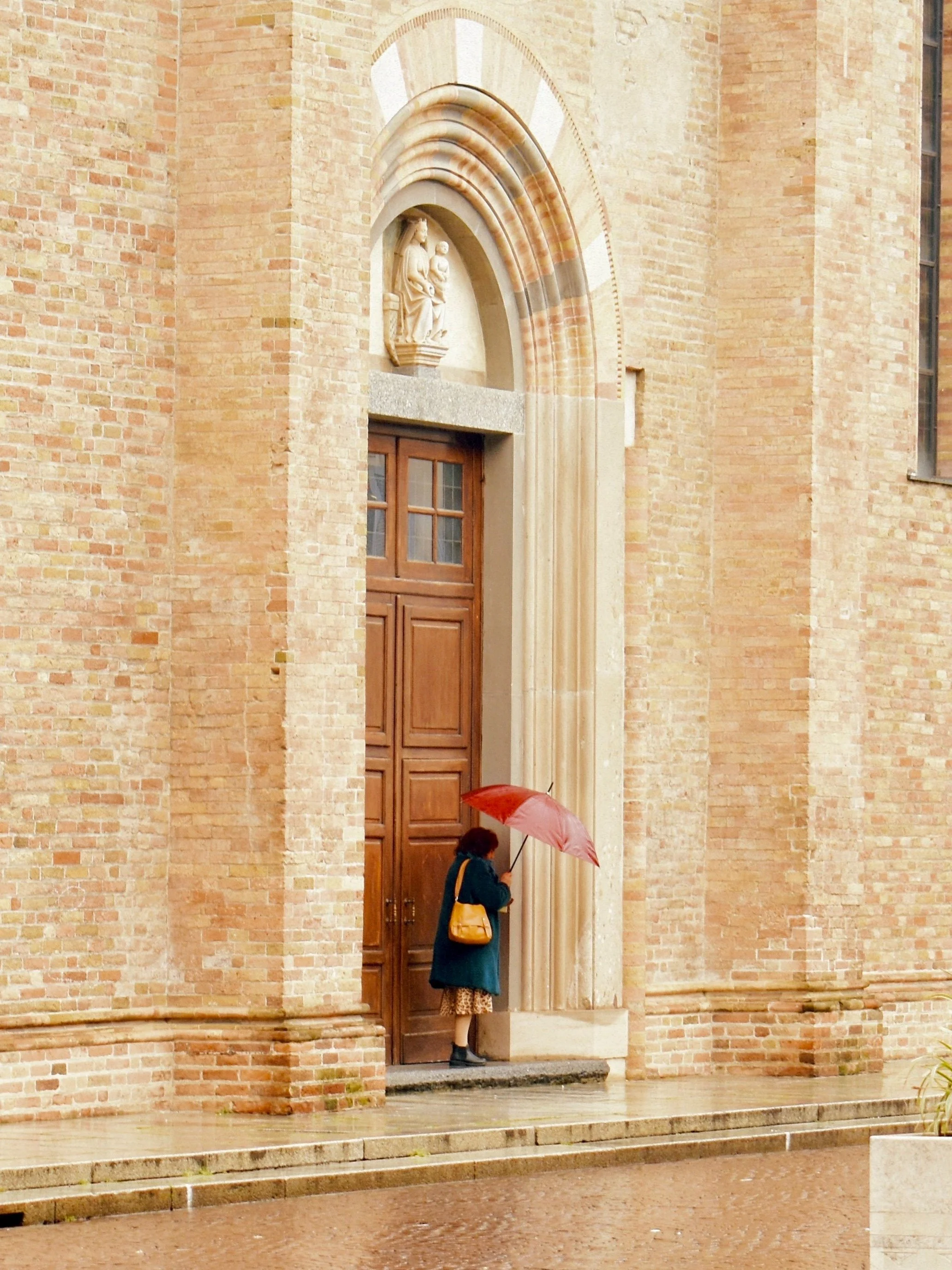 A woman with a yellow purse holding a pink umbrella standing at the entrance of a brick church under a religious sculpture above the door.