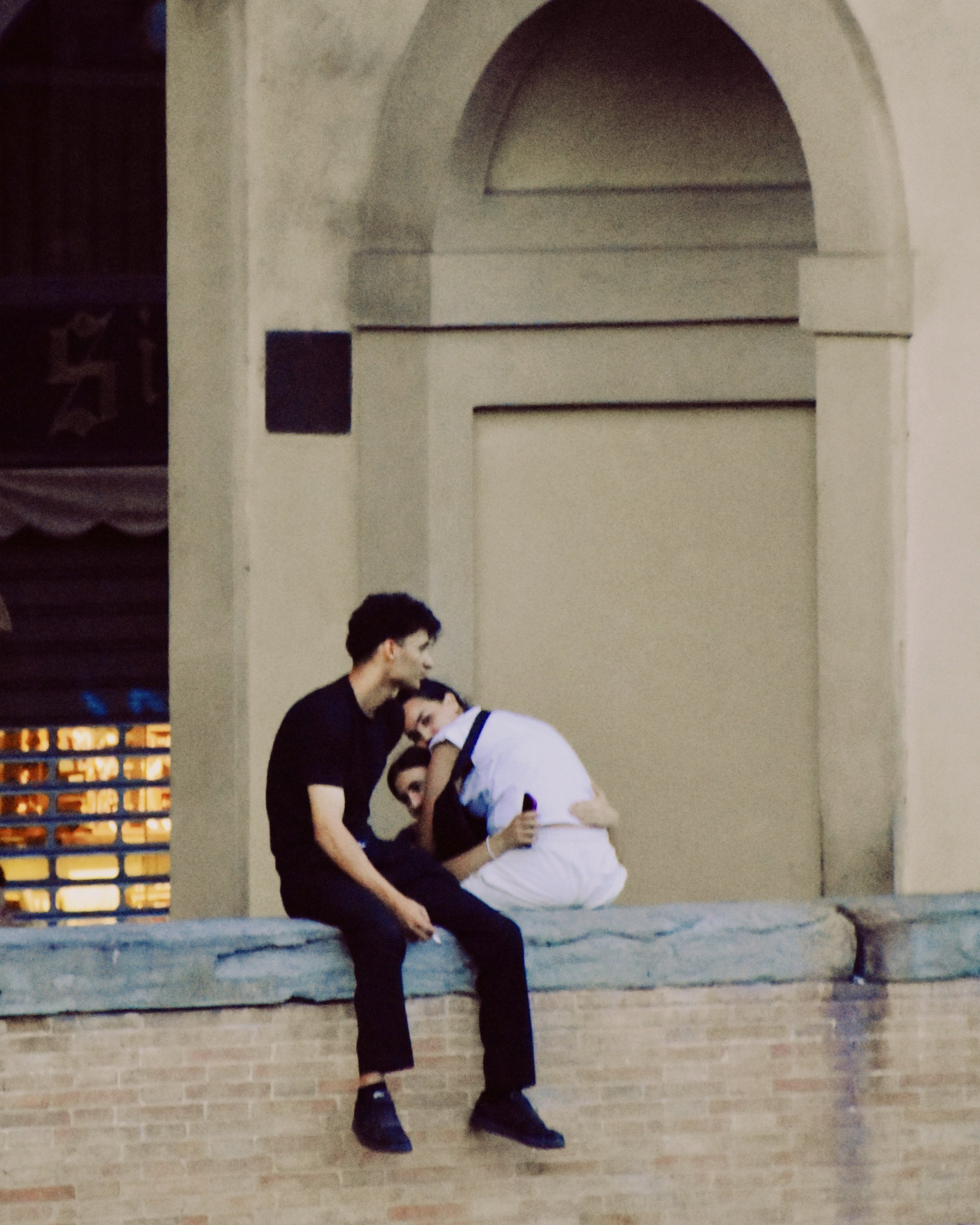 Two men sit on a ledge and affectionately hug each other, smiling, with one holding a phone. They are outdoors in front of a building with an arched window.