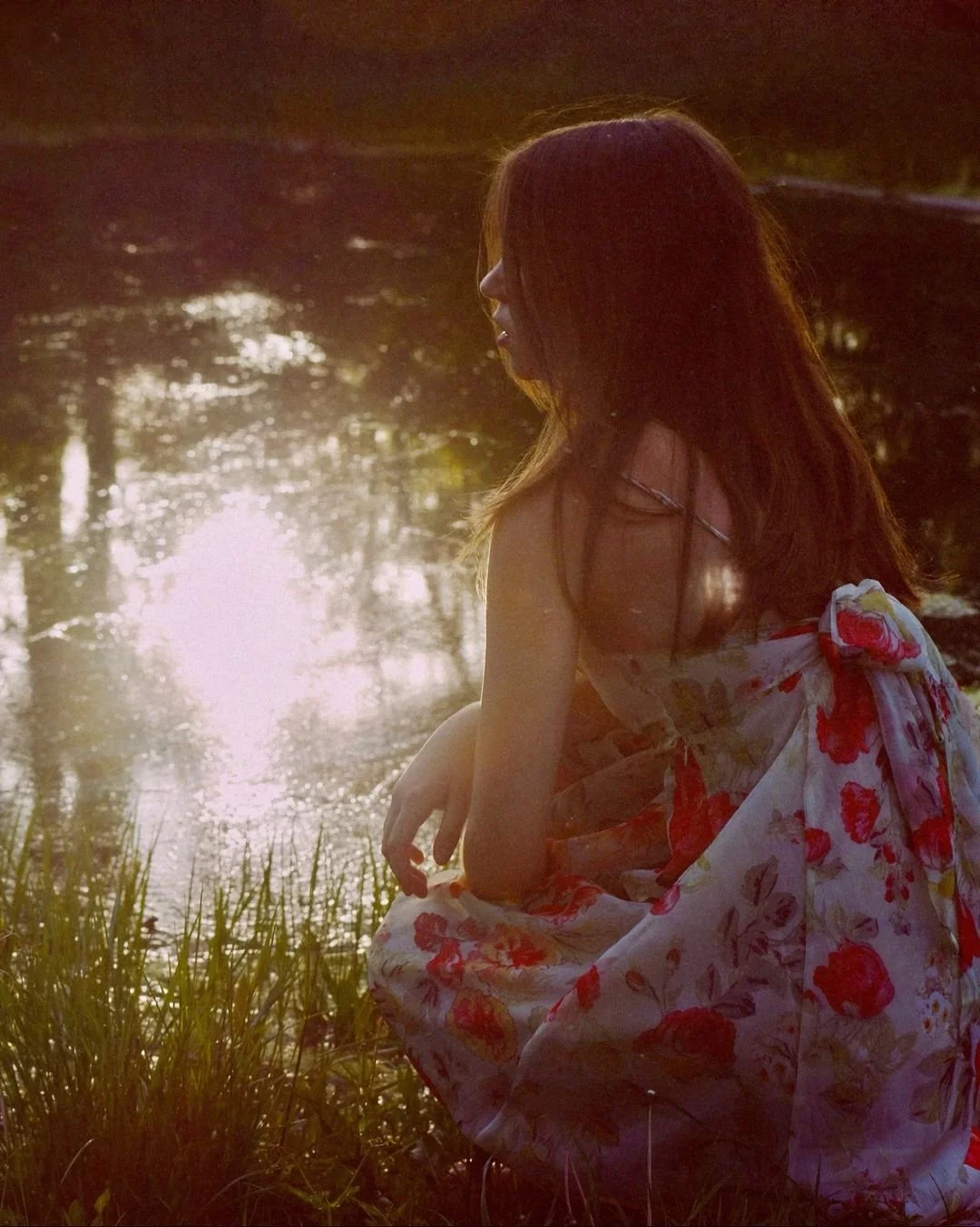 A woman with long hair sitting by a pond or small lake during sunset, wearing a floral dress, with sunlight reflecting on the water.