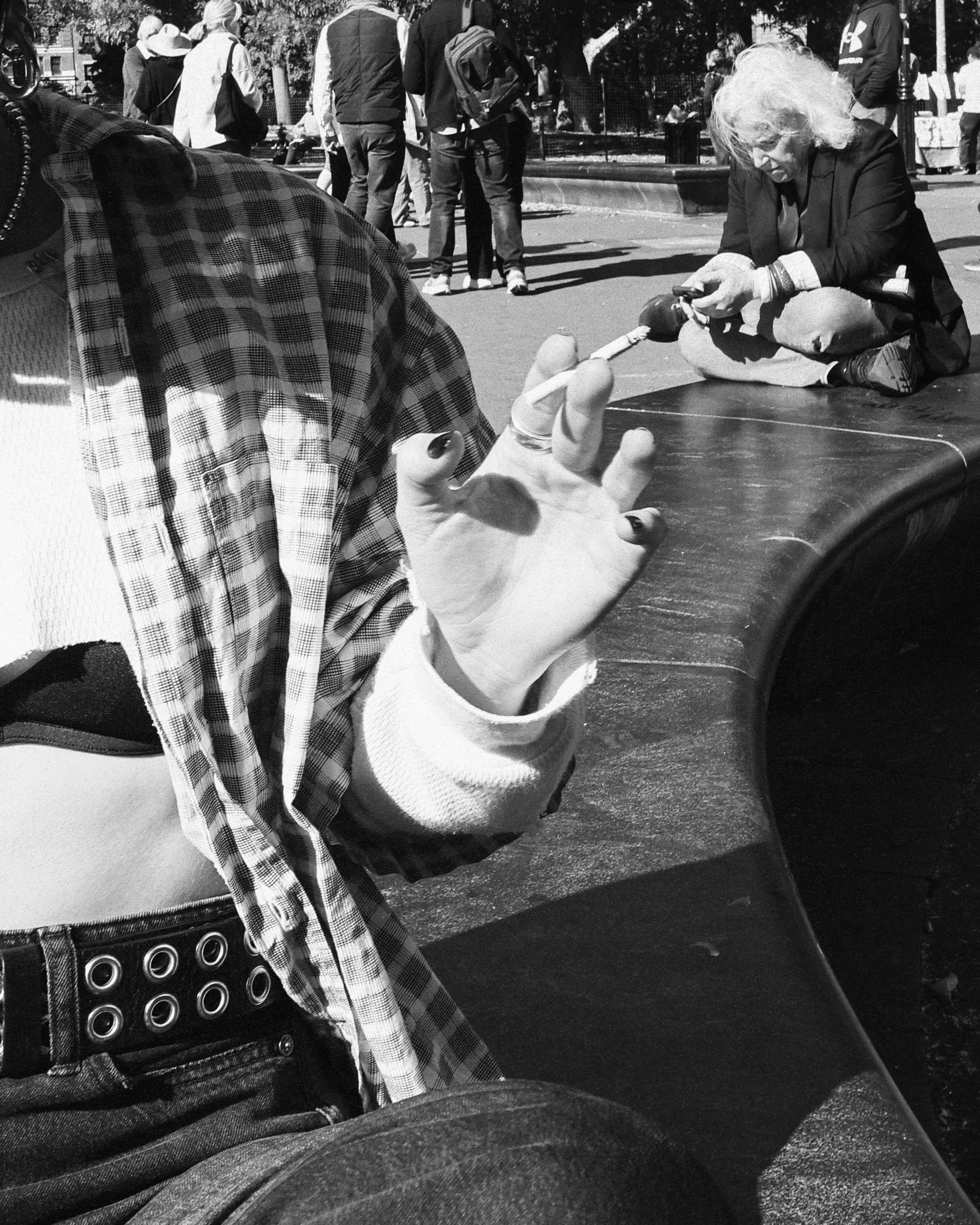 A person makes a peace sign with their hand in the foreground, with an outdoor public space scene in the background. There are multiple people, some standing and some sitting, and a woman with white hair is sitting on the ground, using her phone.