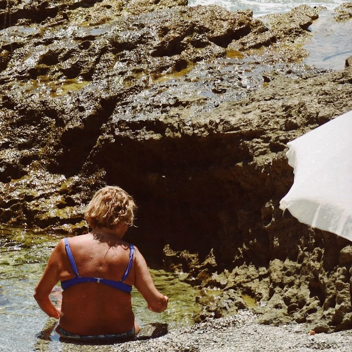 Older woman with short hair, wearing a blue bikini top, sitting in shallow water near rocky shoreline, with rocks and a white umbrella nearby.