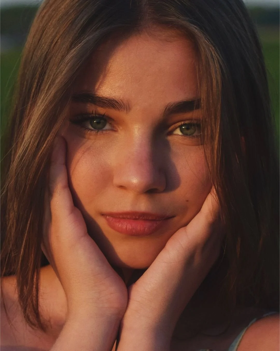 Close-up of a young woman with long brown hair and green eyes, smiling softly, with her hands gently touching her cheeks, captured during sunset.