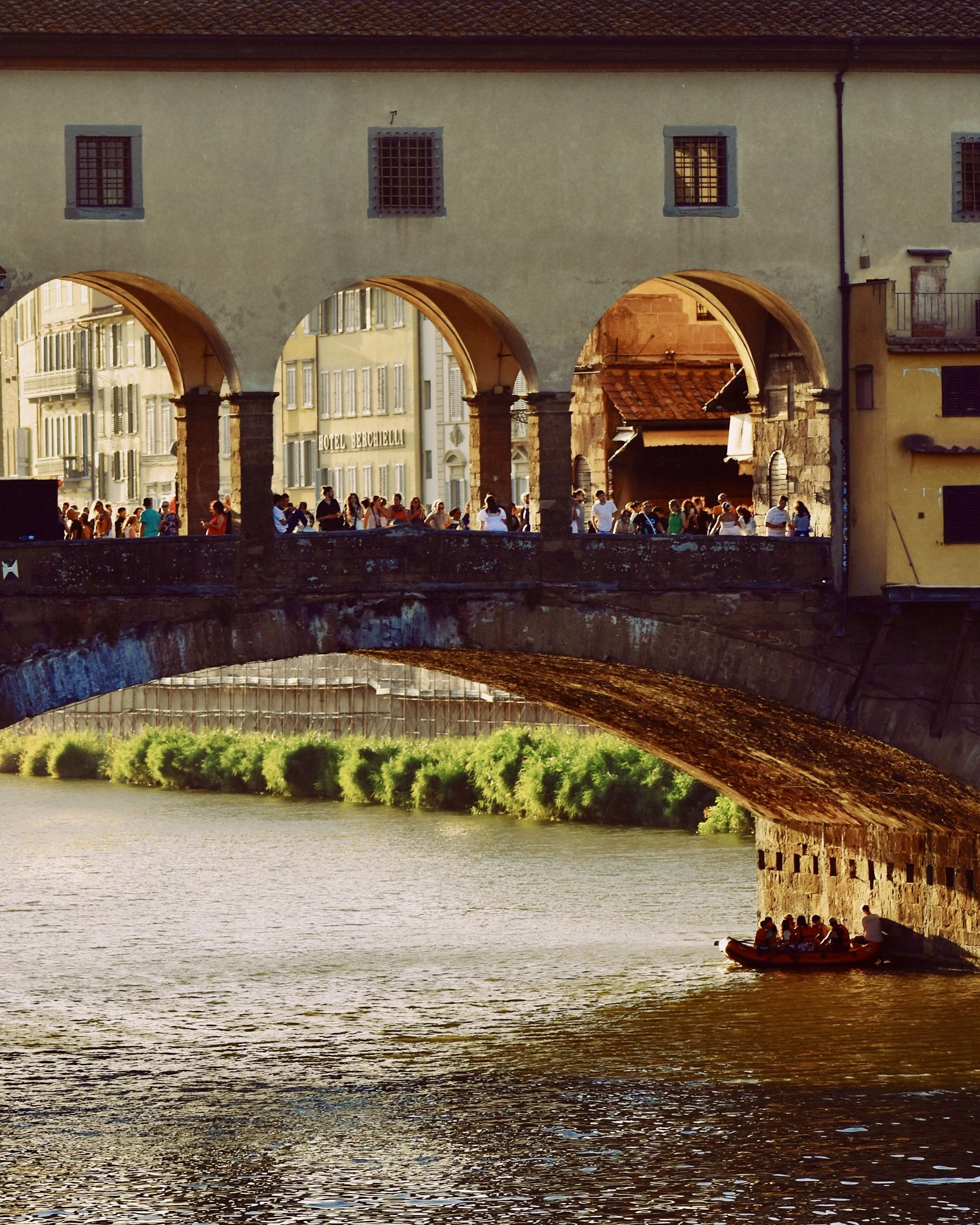 A historic bridge with arches over a river, with a group of tourists in a boat underneath. The bridge has a connect between buildings, and there are pedestrians walking along the street on the bridge. The scene is illuminated by warm sunlight.