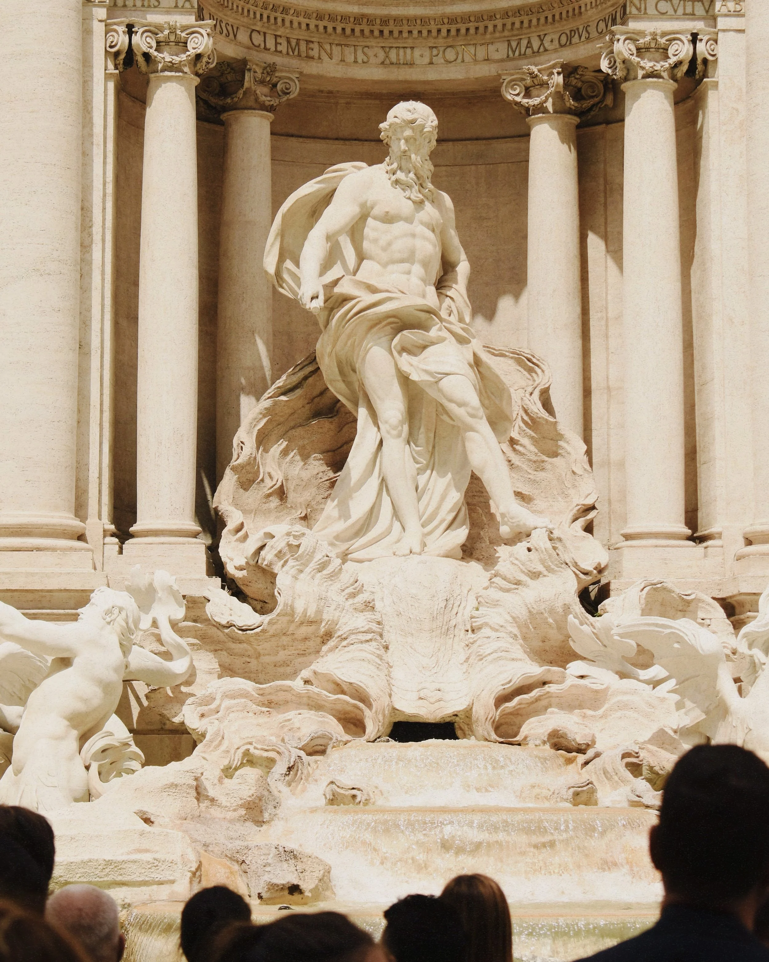 A marble sculpture of a muscular man with a beard, partially draped in cloth, standing atop a rocky base in a colonnade setting. Several waves and mythical figures are at the base, with columns and an inscribed arch in the background.