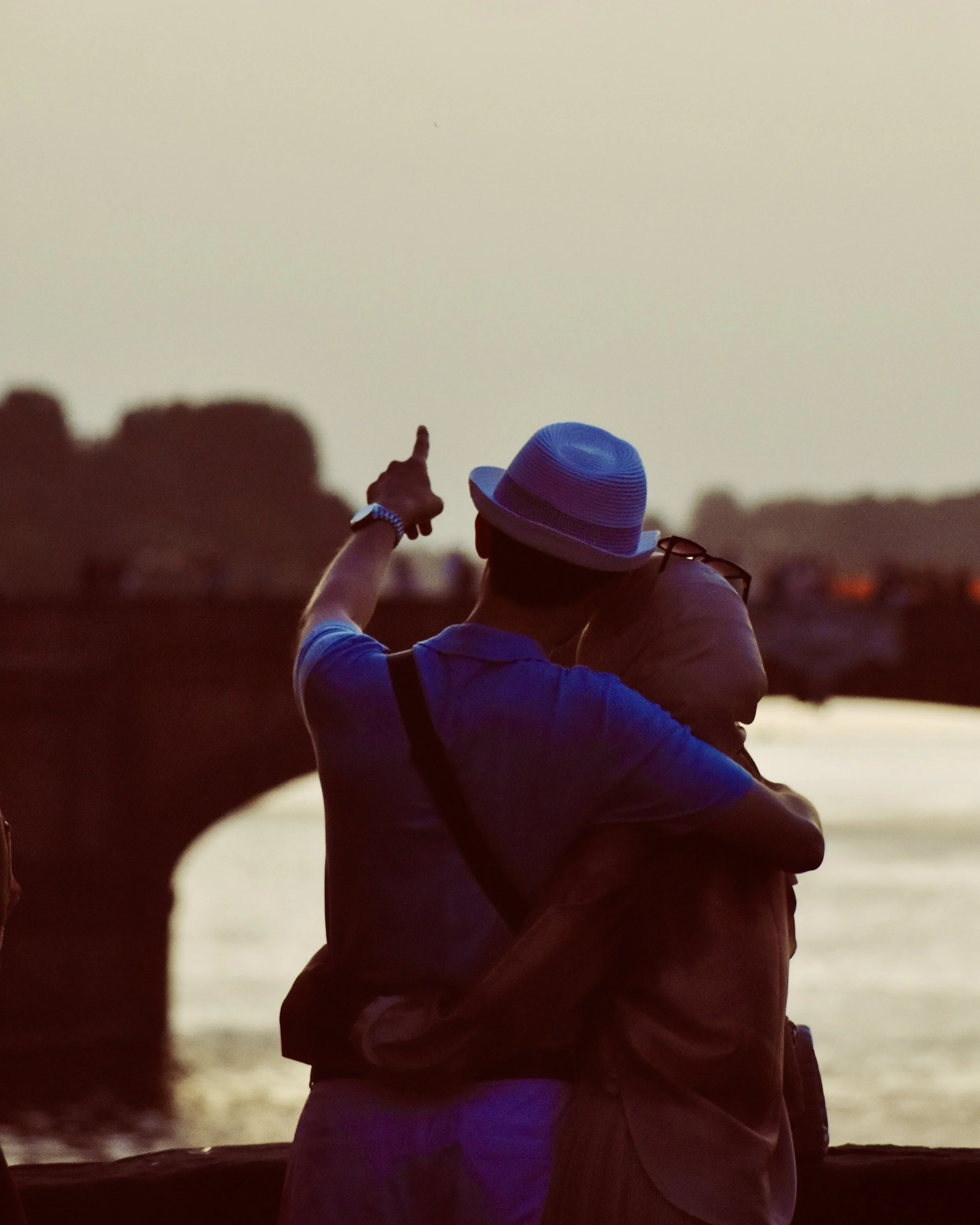 A person wearing a white shirt, a hat, and glasses hugs another person during sunset near the water. The person in the background is pointing towards the horizon.