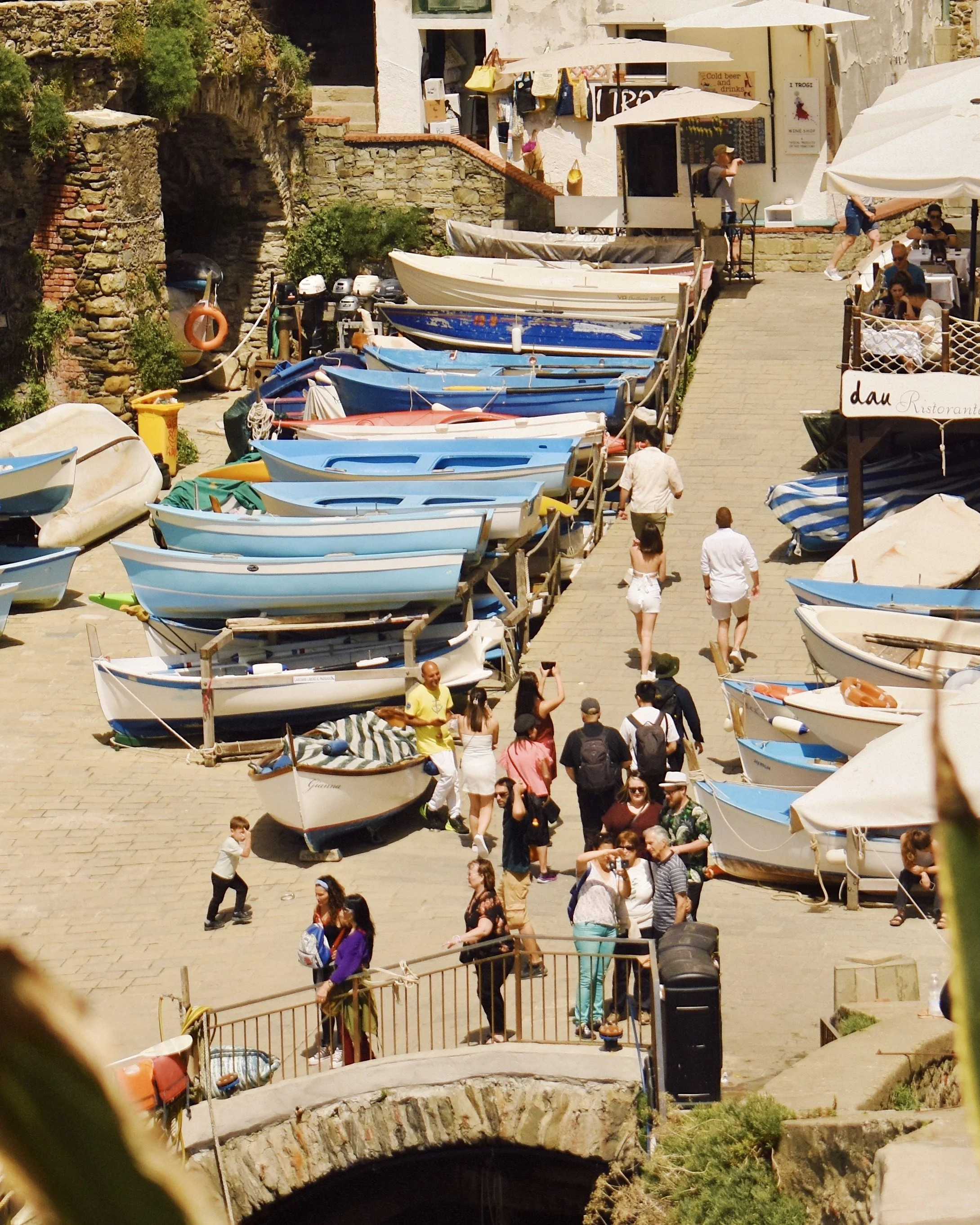 People walking along a docked row of small boats, with outdoor dining and stone buildings in the background.