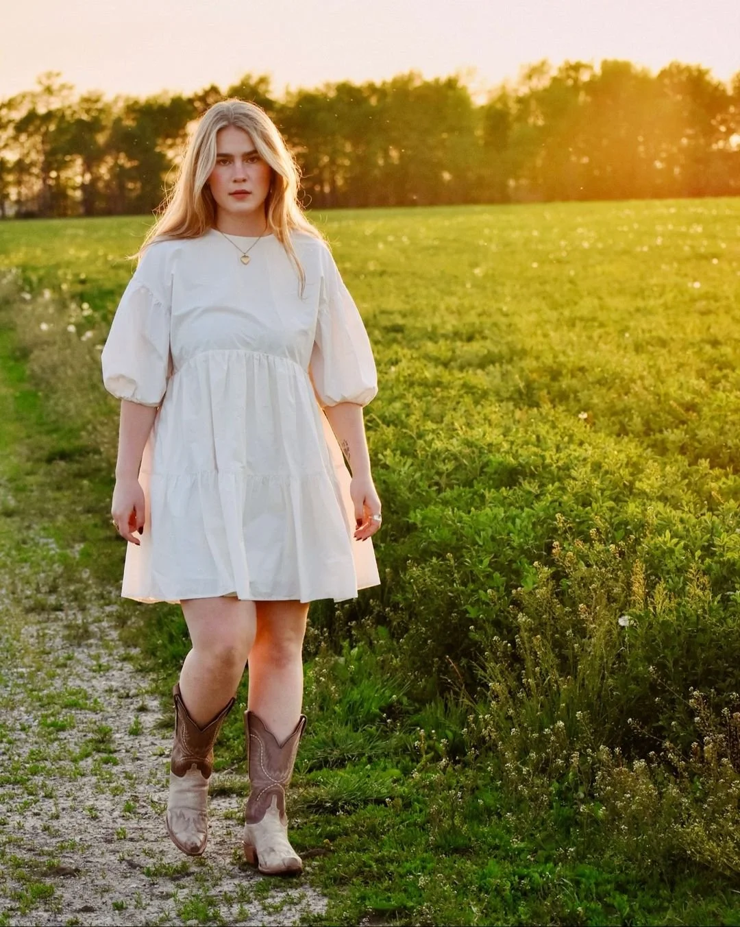 A young woman in a white dress and cowboy boots walking along a rural path at sunset with green fields and trees in the background.