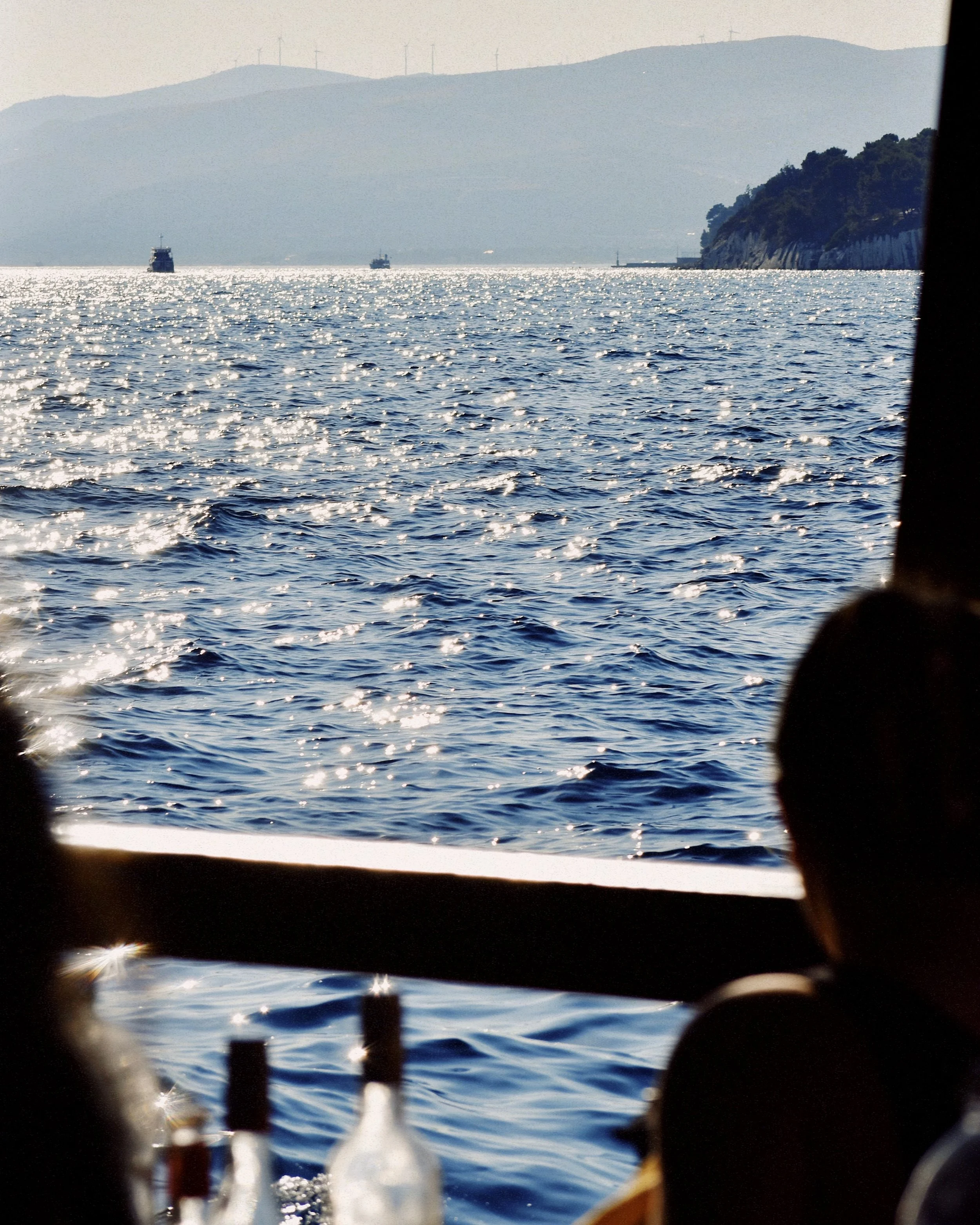 View of the ocean with sunlight reflecting off the water, distant ships, hills, and wind turbines in the background, seen from a boat with partially visible people and bottles in the foreground.