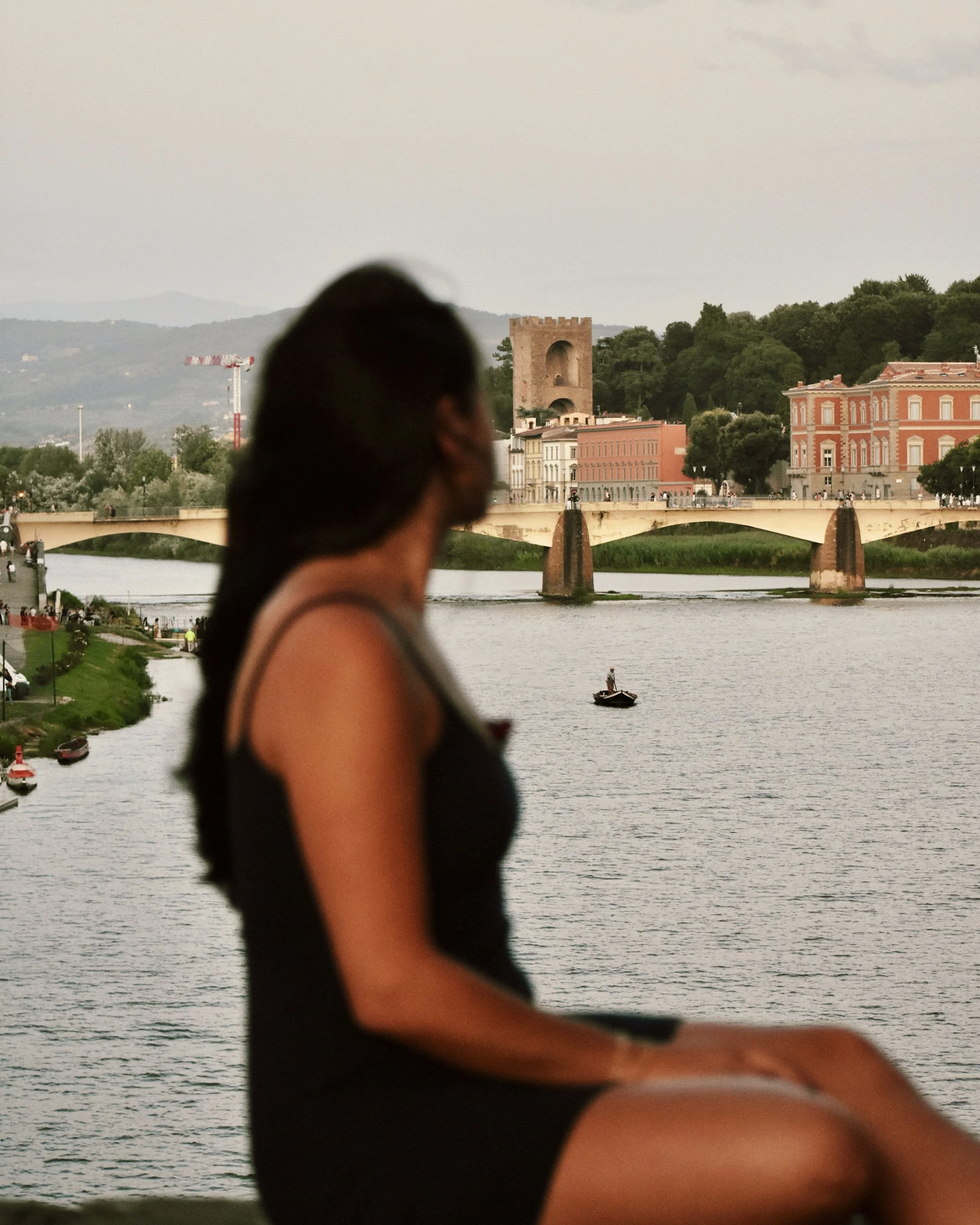 A woman sitting by the water with a cityscape and bridge in the background.