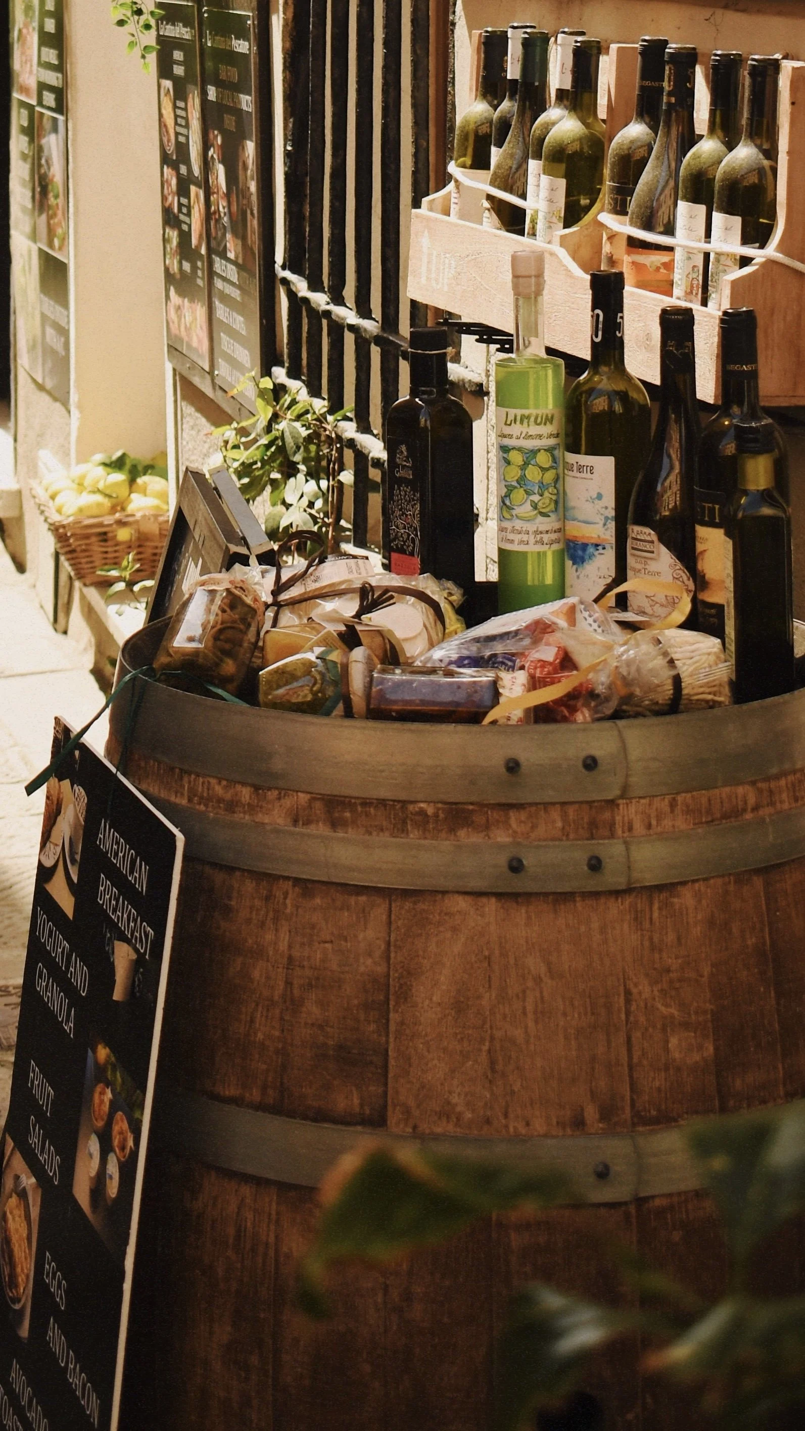 A decorative display of wine bottles, olive oil bottles, and packaged food items arranged on a large wooden barrel outside a store, with a blackboard sign advertising American breakfast options.