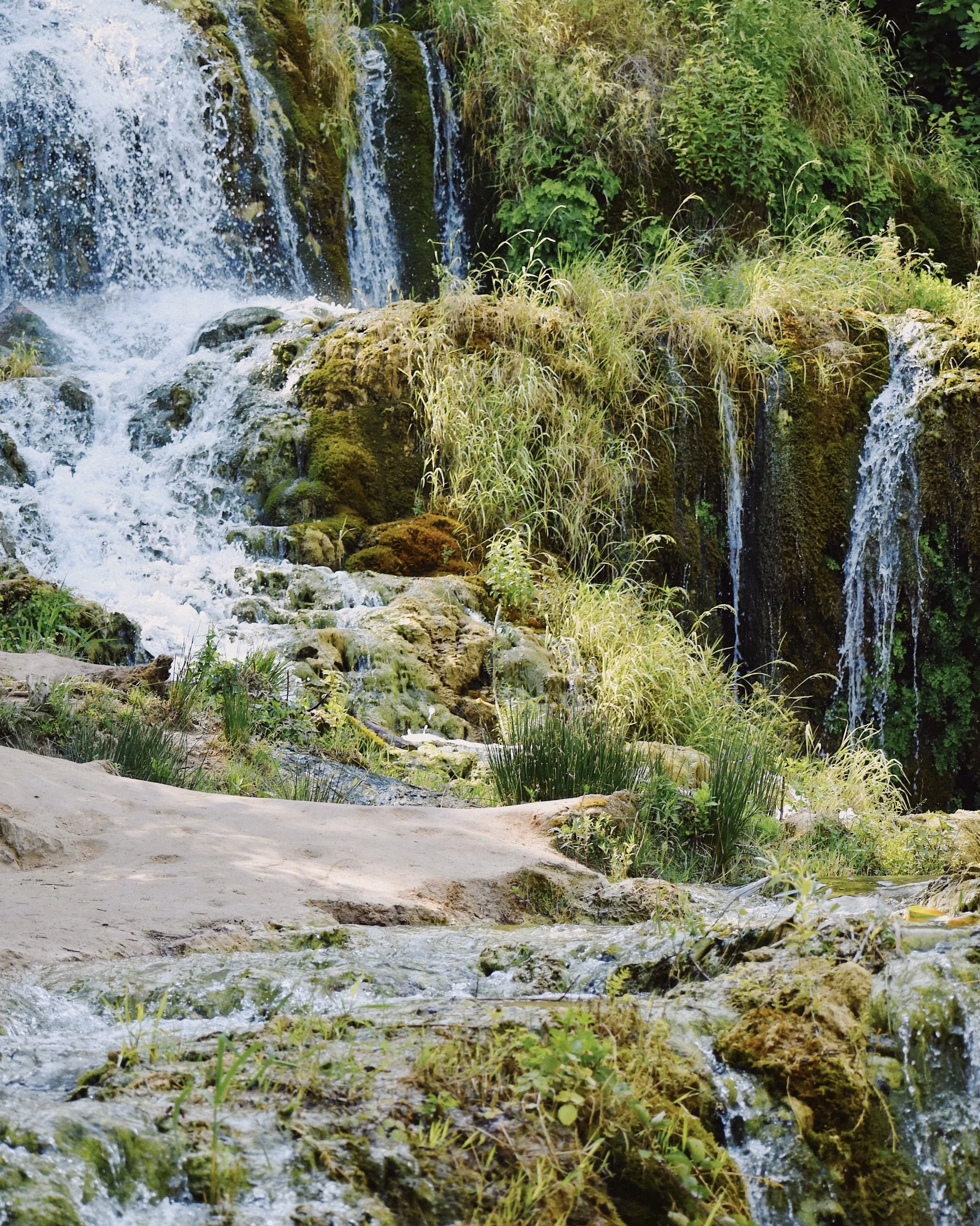 A scenic view of a small waterfall with water cascading over rocks, surrounded by green grass and lush vegetation.