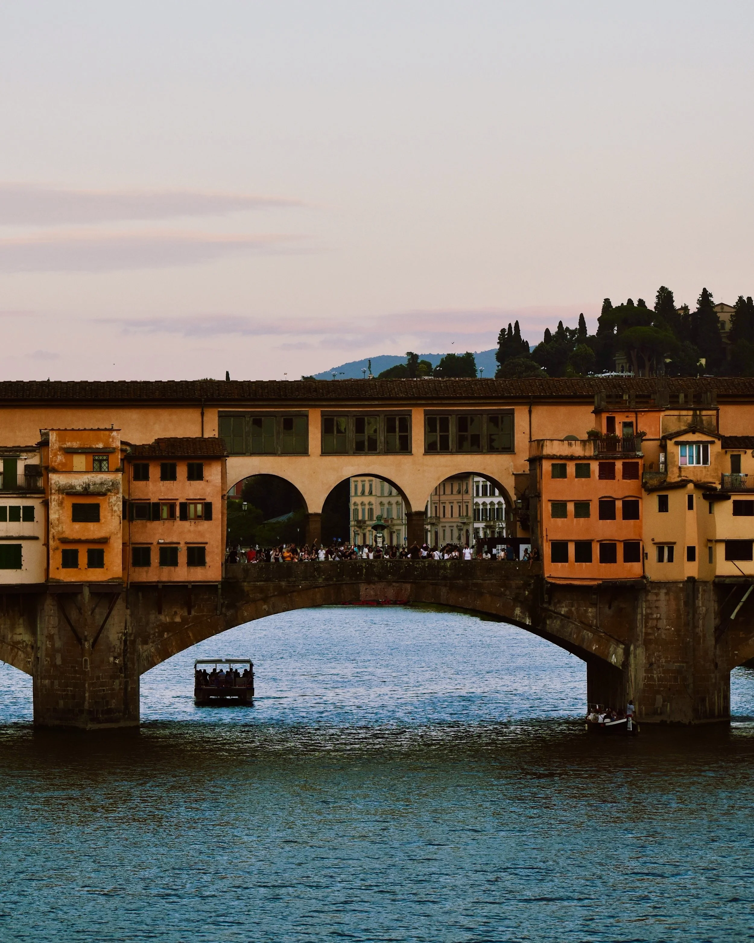 View of the Ponte Vecchio bridge over the Arno River in Florence, Italy, with buildings and people on the bridge and hills in the background during sunset.