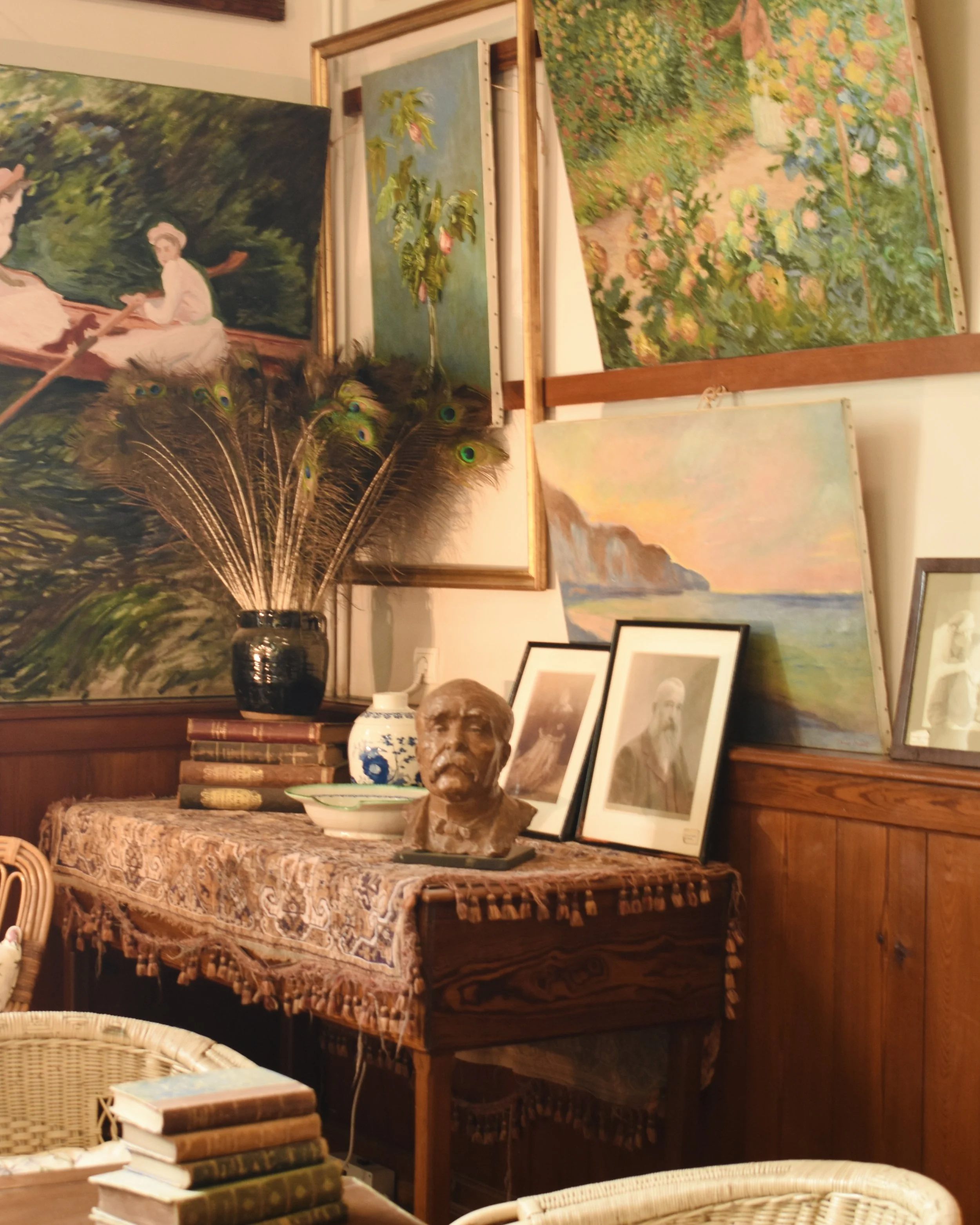 An art gallery room decorated with various paintings, a table covered with a decorative cloth, a vase with peacock feathers, and framed photographs and sculptures.